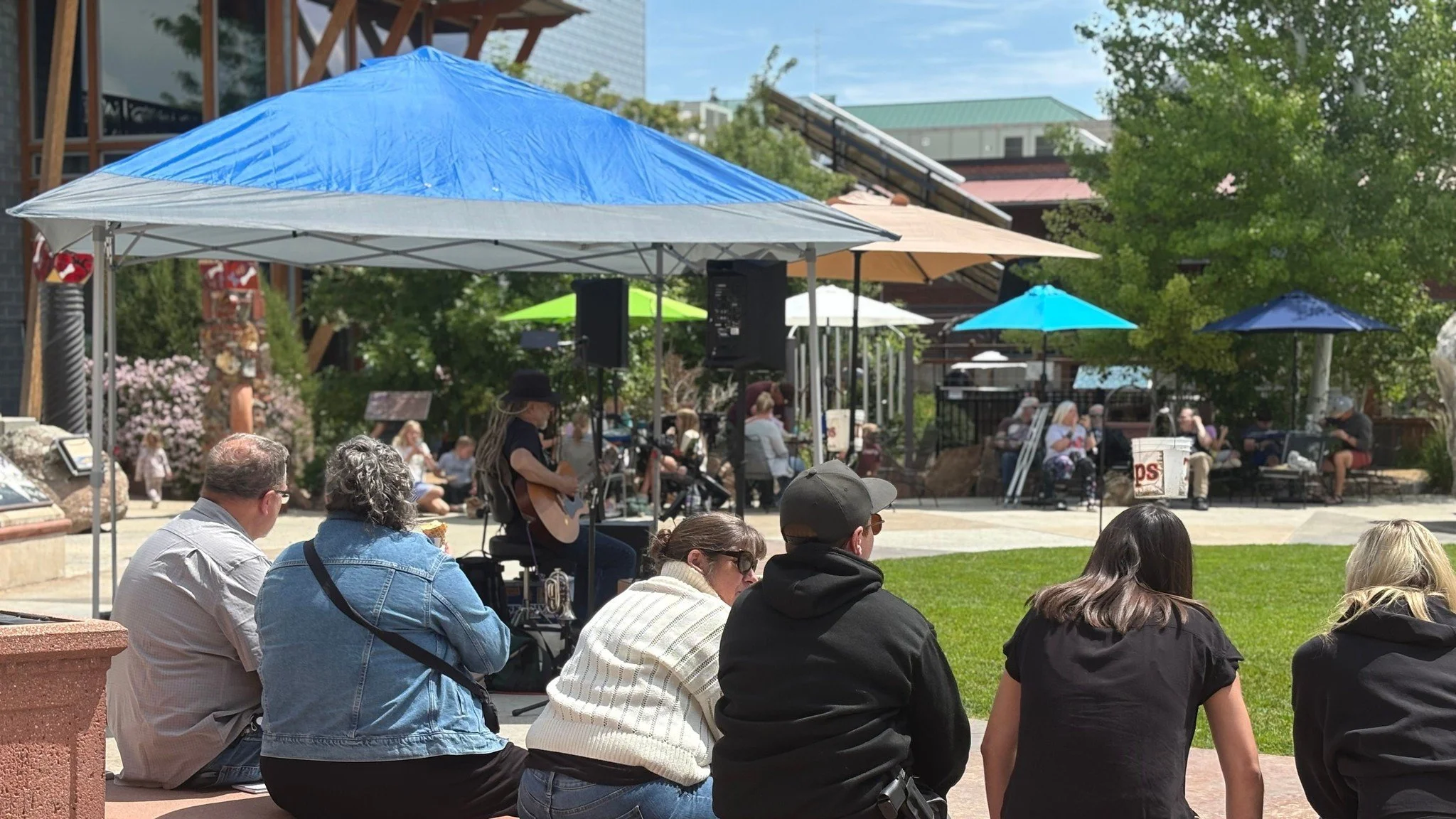 People sitting on steps and lawn, watching a street performer playing guitar and singing under umbrellas, in a park-like area with trees and buildings in the background.