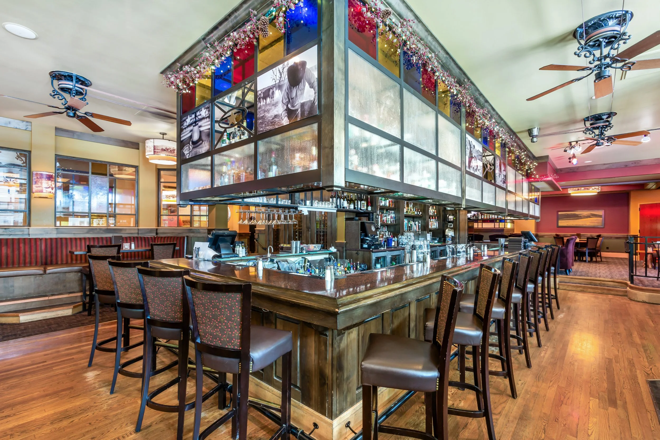 A restaurant bar with a large, curved wooden counter, high chairs, and a modern, illuminated overhead shelf decorated with fairy lights and photos. The interior features a high ceiling with ceiling fans, large windows, and warm lighting.