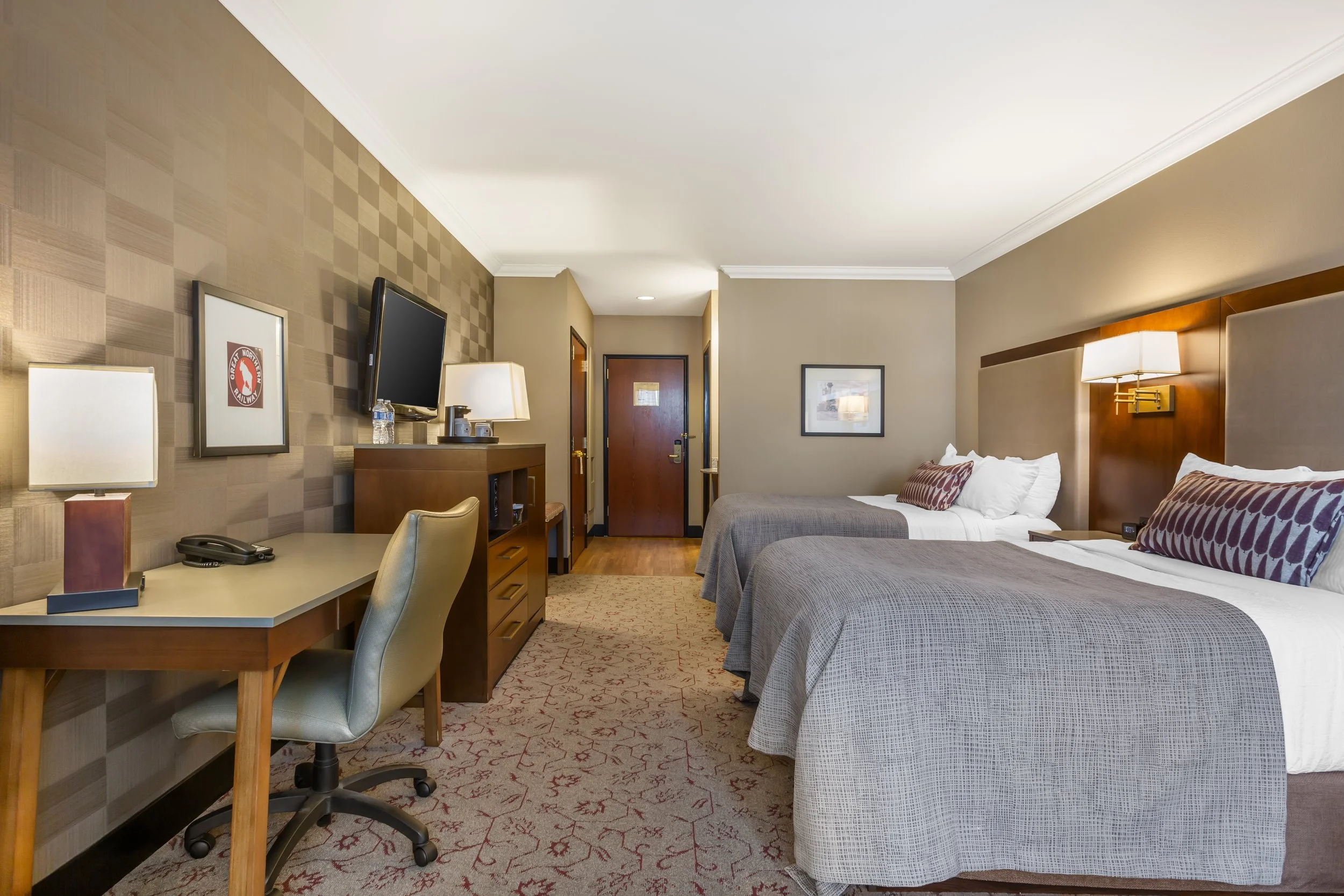 Hotel room with two queen beds, a wooden desk with a beige swivel chair, a wall-mounted TV, lamps, and artwork on the walls.
