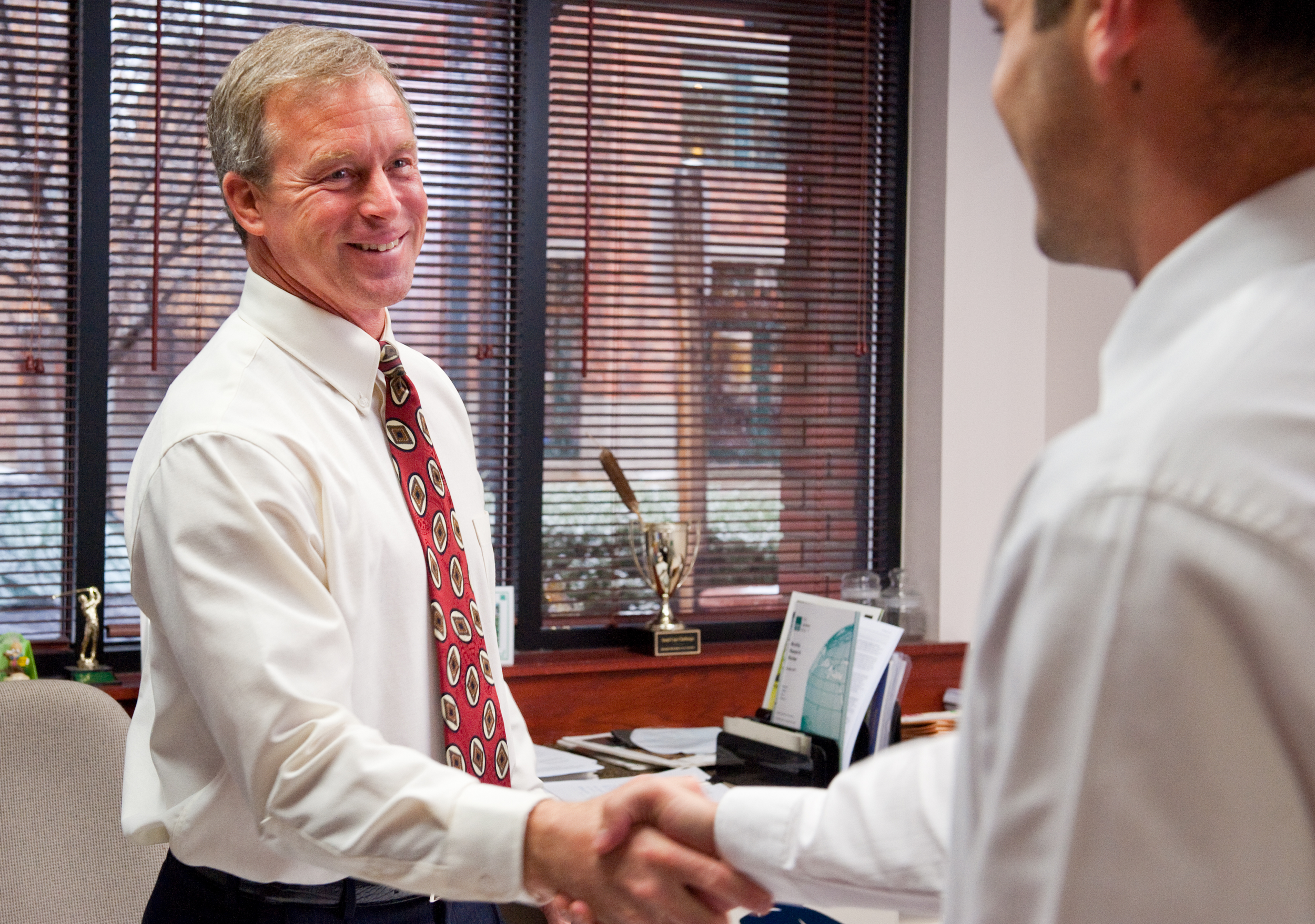 Two businessmen shaking hands in an office, smiling. One man has gray hair and a patterned tie, the other has short brown hair. The office has a desk with papers, a trophy, and a window with blinds.