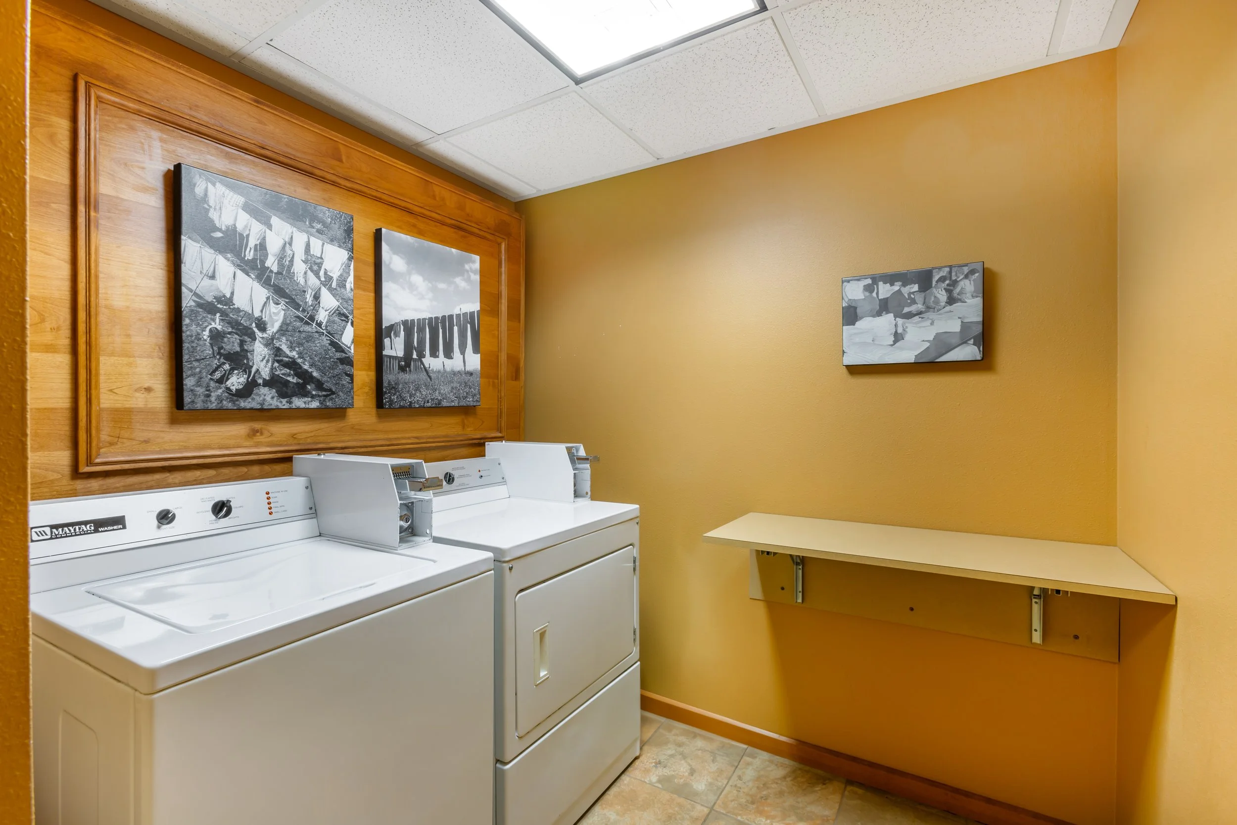 A laundry room with a white Maytag washing machine and dryer, a wooden framed wall with black-and-white photos of laundry and clothes hung outside, a yellow wall, and a fold-out table on the right.