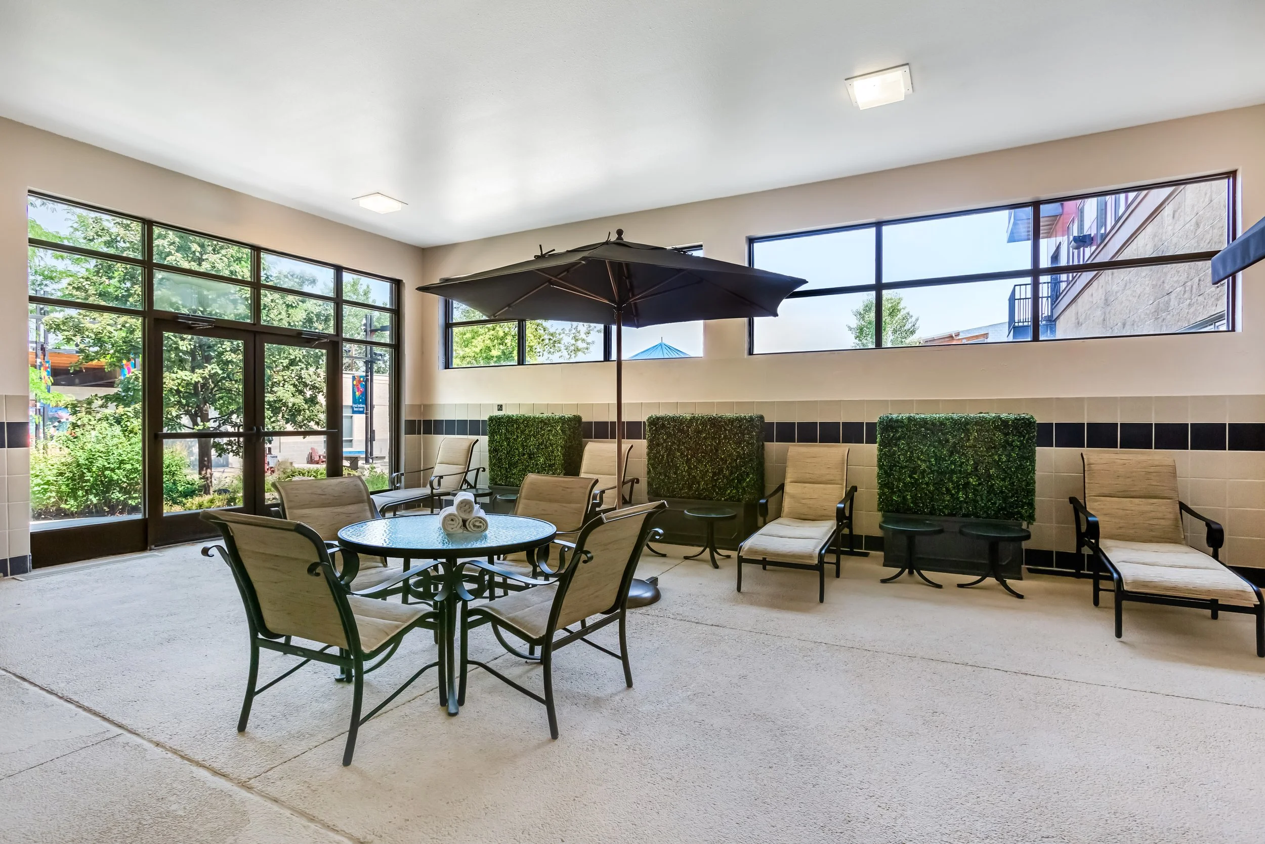 Indoor patio area with chairs, a round glass table with rolled towels, a large umbrella, and green hedges in planters. Large windows let in natural light.