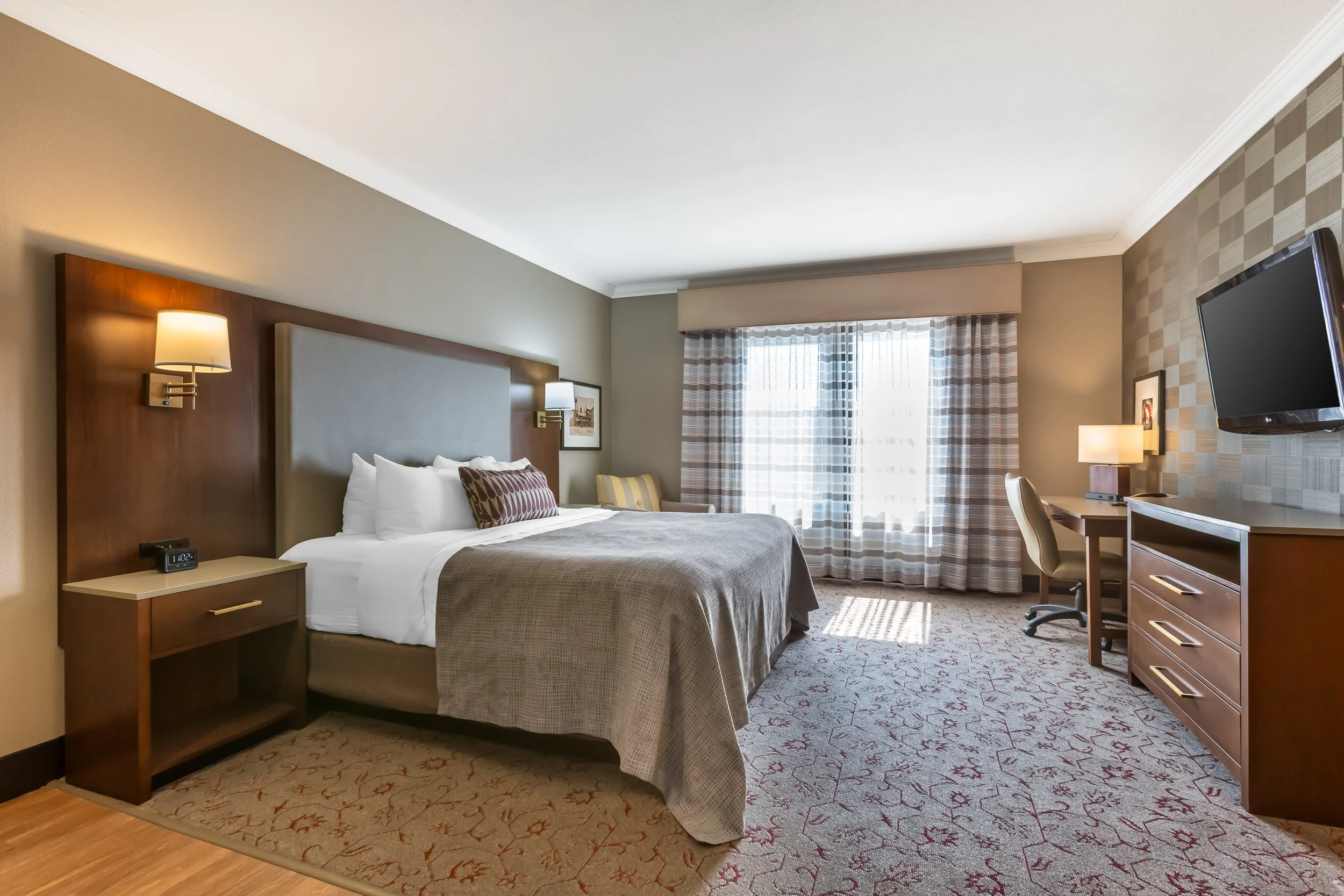 A hotel room with a large bed, nightstands, wall-mounted lamps, a flat-screen TV, a desk, a chair, and a window with curtains.