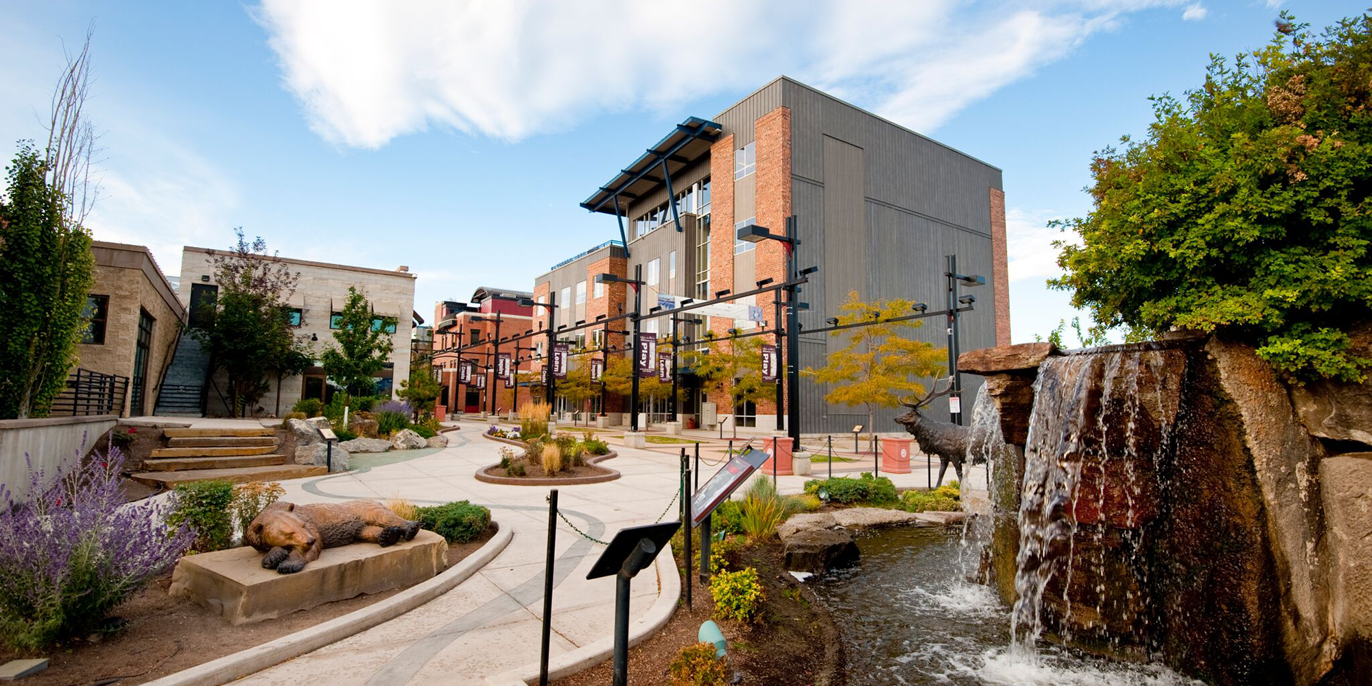 An outdoor urban area featuring a modern multi-story building, water fountain with waterfall, sculptures of animals including a dog and a bear, trees, plants, and a pathway with informational signs.