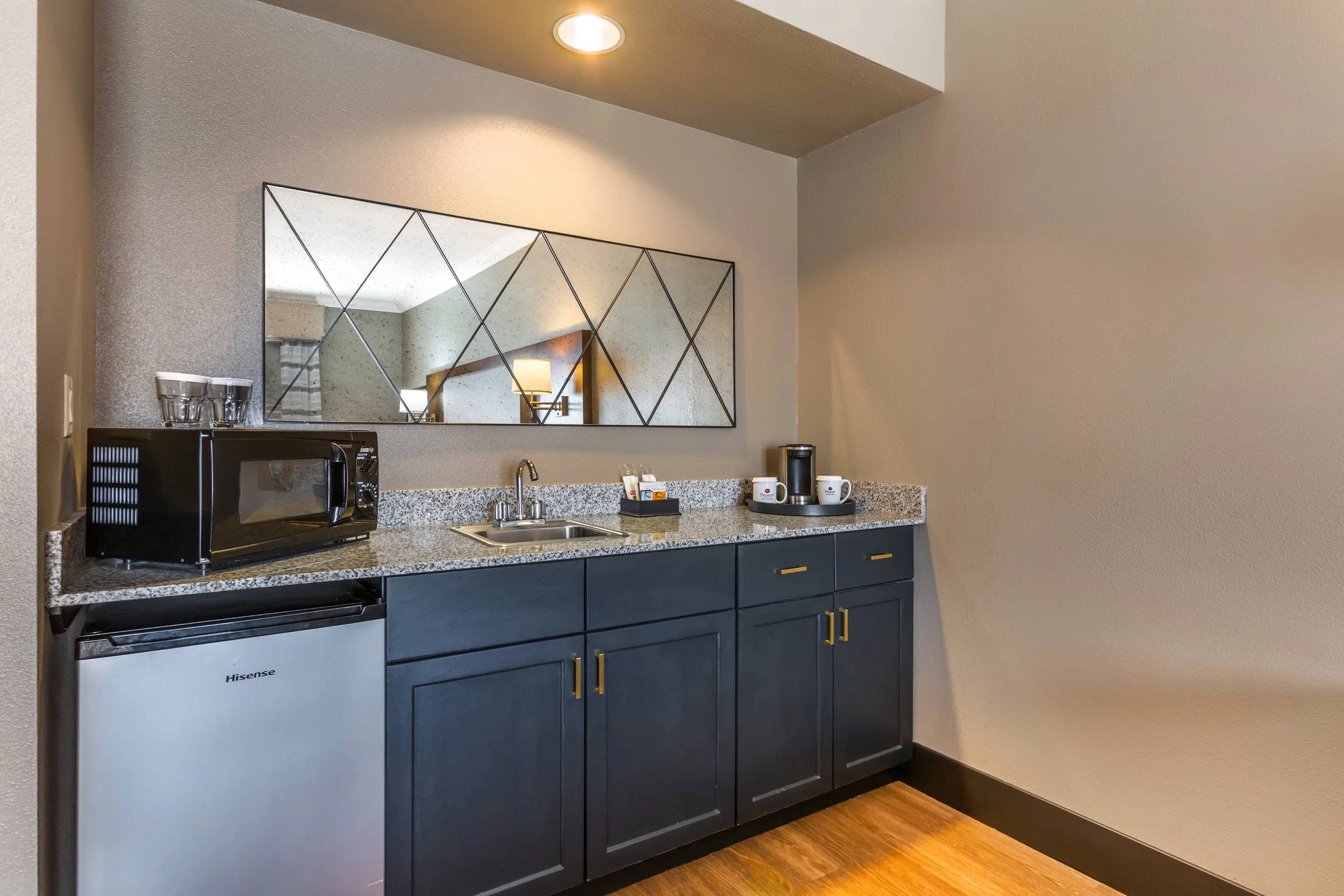 Mini fridge, microwave, coffee maker, cups, and supplies on kitchen countertop with mirror and wall light reflection