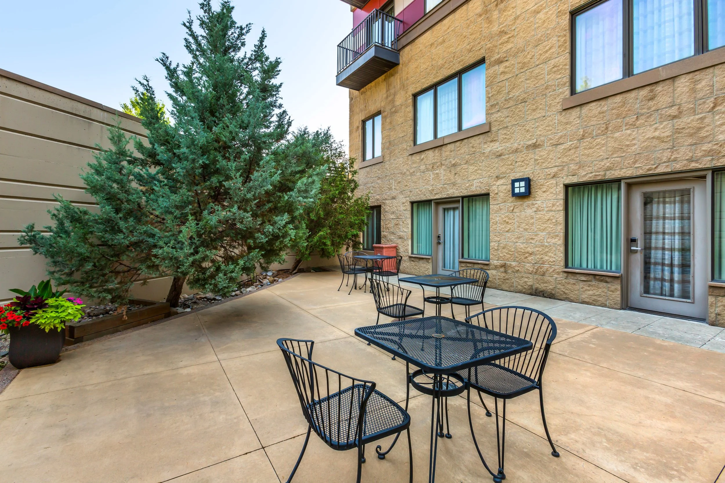 Outdoor patio area with black metal tables and chairs, a potted plant with colorful flowers, large trees, and an apartment building with beige brick walls and windows.