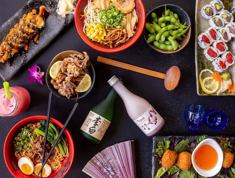 Assorted Japanese dishes on a black table, including sushi rolls, ramen, edamame, fried calamari, a sushi roll with eel, a bowl of noodles with boiled eggs, a bottle of sake, and various drinks.