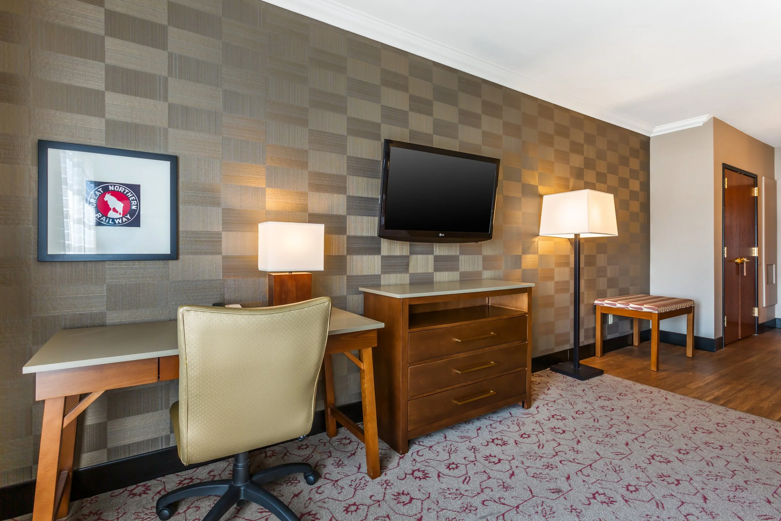 Interior of a hotel room with a desk and chair, a dresser with a mounted TV, two lamps, a framed wall art, a bench, and a doorway, with a patterned carpet and wooden flooring.