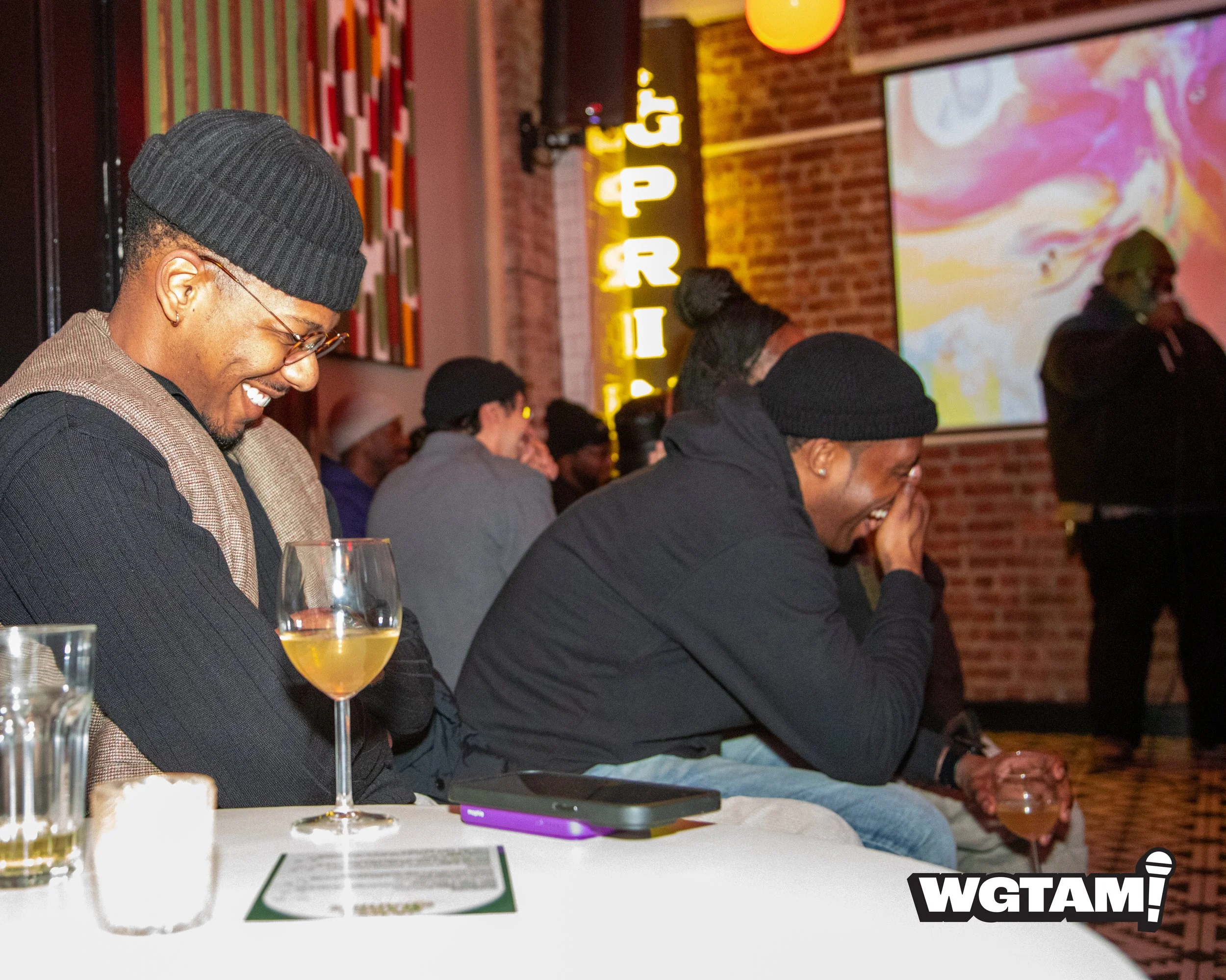 Two men are sitting at a table in a dimly lit venue, both laughing and enjoying drinks. The man on the left is wearing a black beanie, glasses, and a checked vest, while the man on the right is wearing a black beanie and black hoodie. There are several glasses and a menu on the table, and other people are visible in the background, some also laughing. The background features colorful artwork, a brick wall, and a neon sign.
