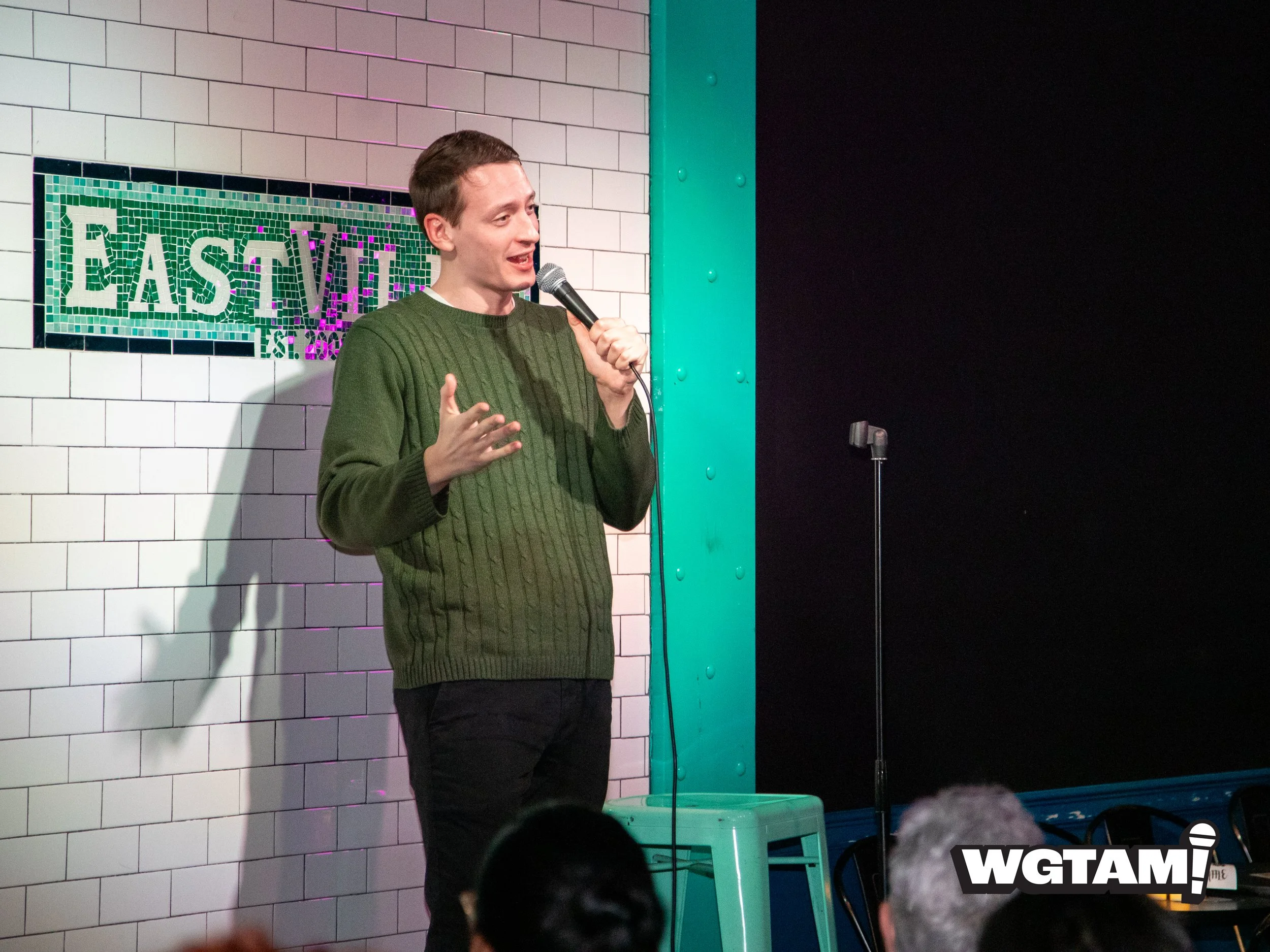 A man with short brown hair and a green sweater is holding a microphone and speaking on stage at a comedy club or performance venue. The background features a white tiled brick wall, a mosaic sign that reads "EAST V" partially visible, and a teal-colored structural element. There is an empty teal stool next to him, and an audience member with gray hair is visible in front. The logo "WGTAM!" is in the bottom right corner of the image.