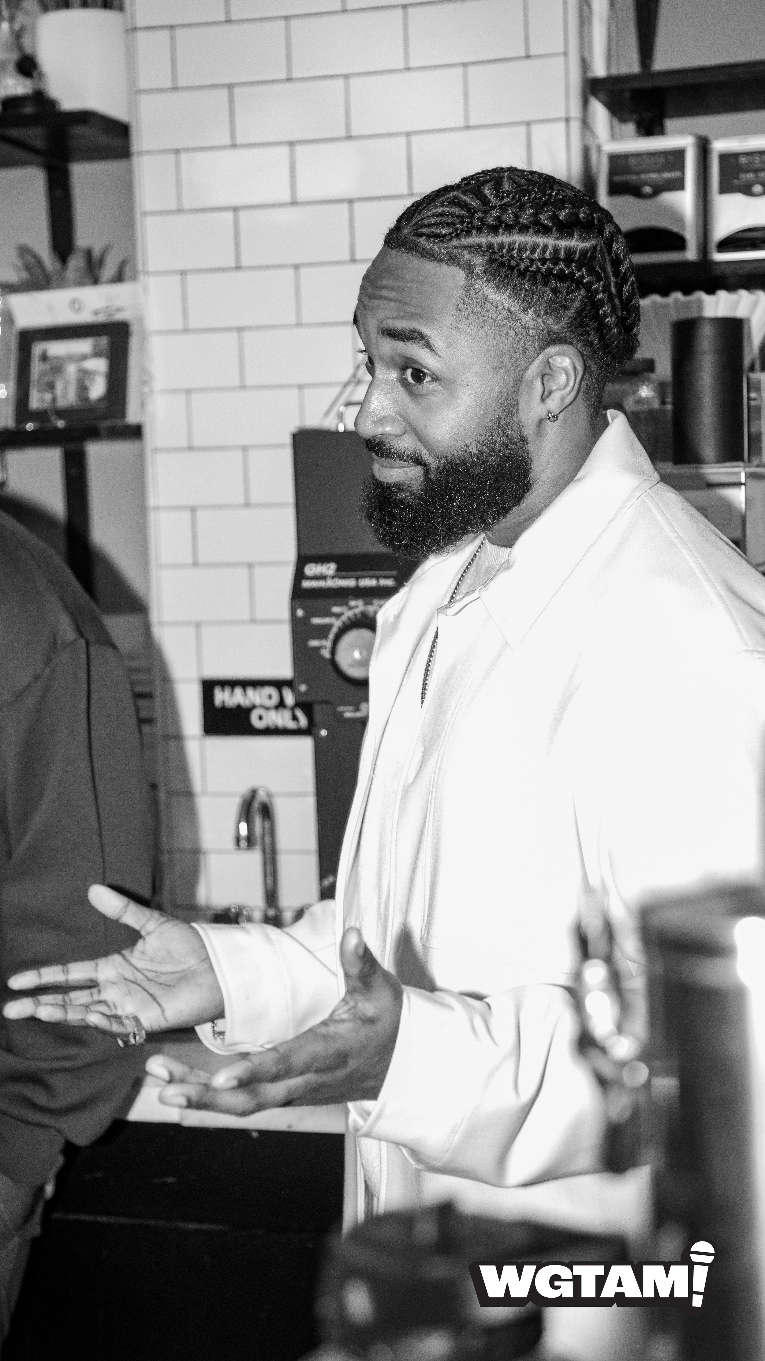 A man with a beard and braided hair, wearing a light-colored shirt, is gesturing with his hands while talking in a cafe or restaurant setting.
