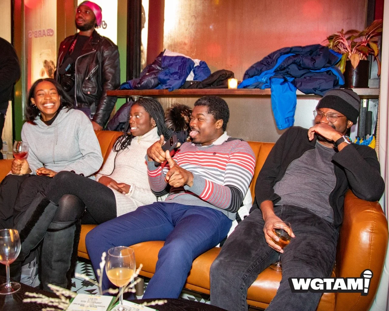 Group of five diverse friends sitting on a brown leather couch at a social gathering, laughing and enjoying drinks in a cozy indoor setting.