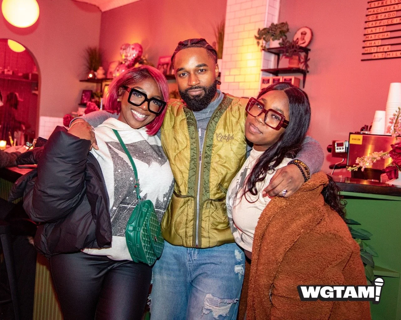 Three people standing closely together at an indoor event, smiling and posing for the camera. The woman on the left has pink hair, wearing glasses, a white hoodie, and a black jacket. The man in the middle has a beard, wearing a yellow jacket and ripped jeans. The woman on the right has glasses, long black hair, and is wearing a brown coat. The background features pink lighting, a bar counter, and decorative shelves.