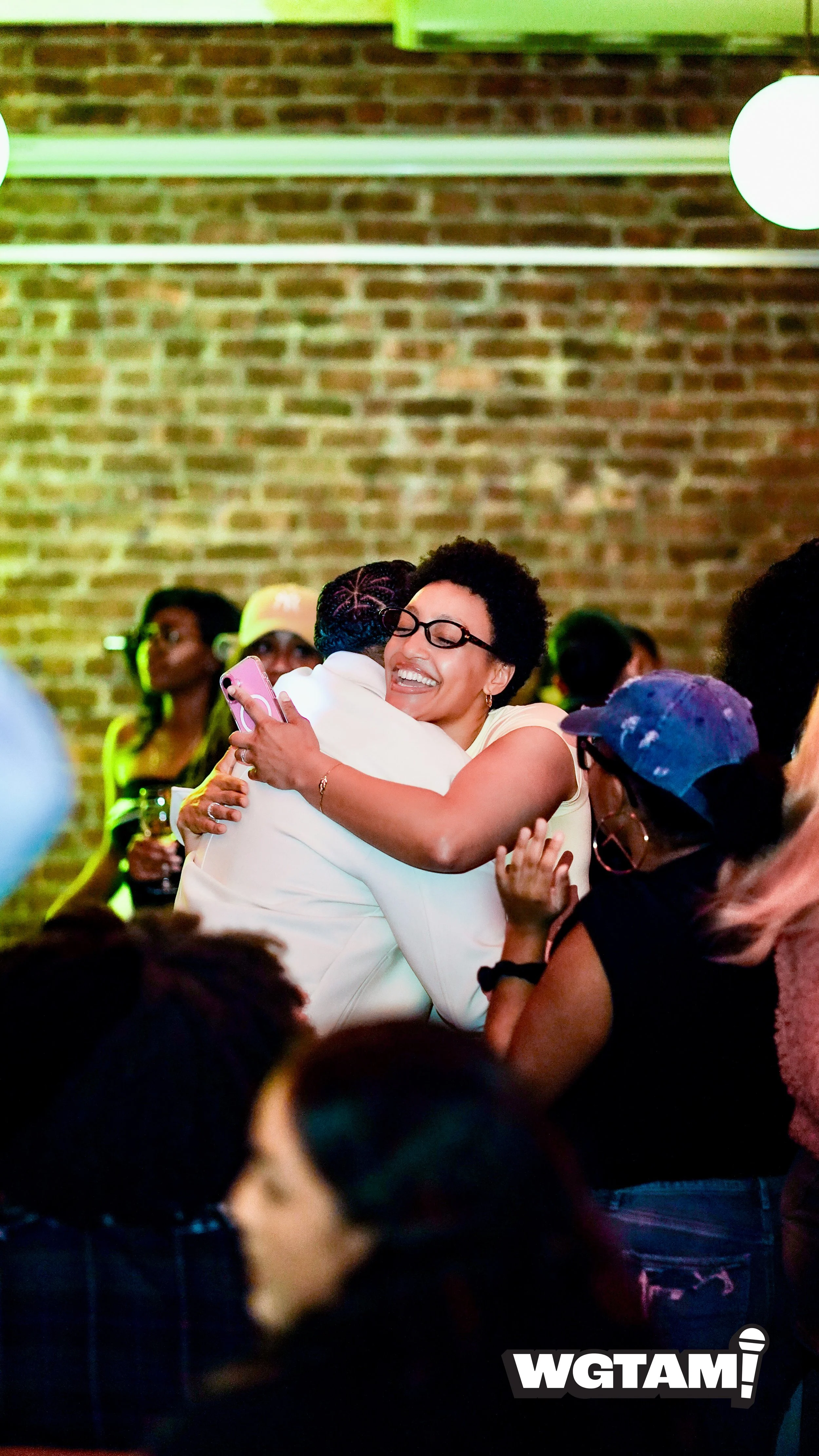 A woman with glasses and short curly hair hugging a person in a white shirt while smiling at a lively social event with a brick wall in the background and people wearing headphones.