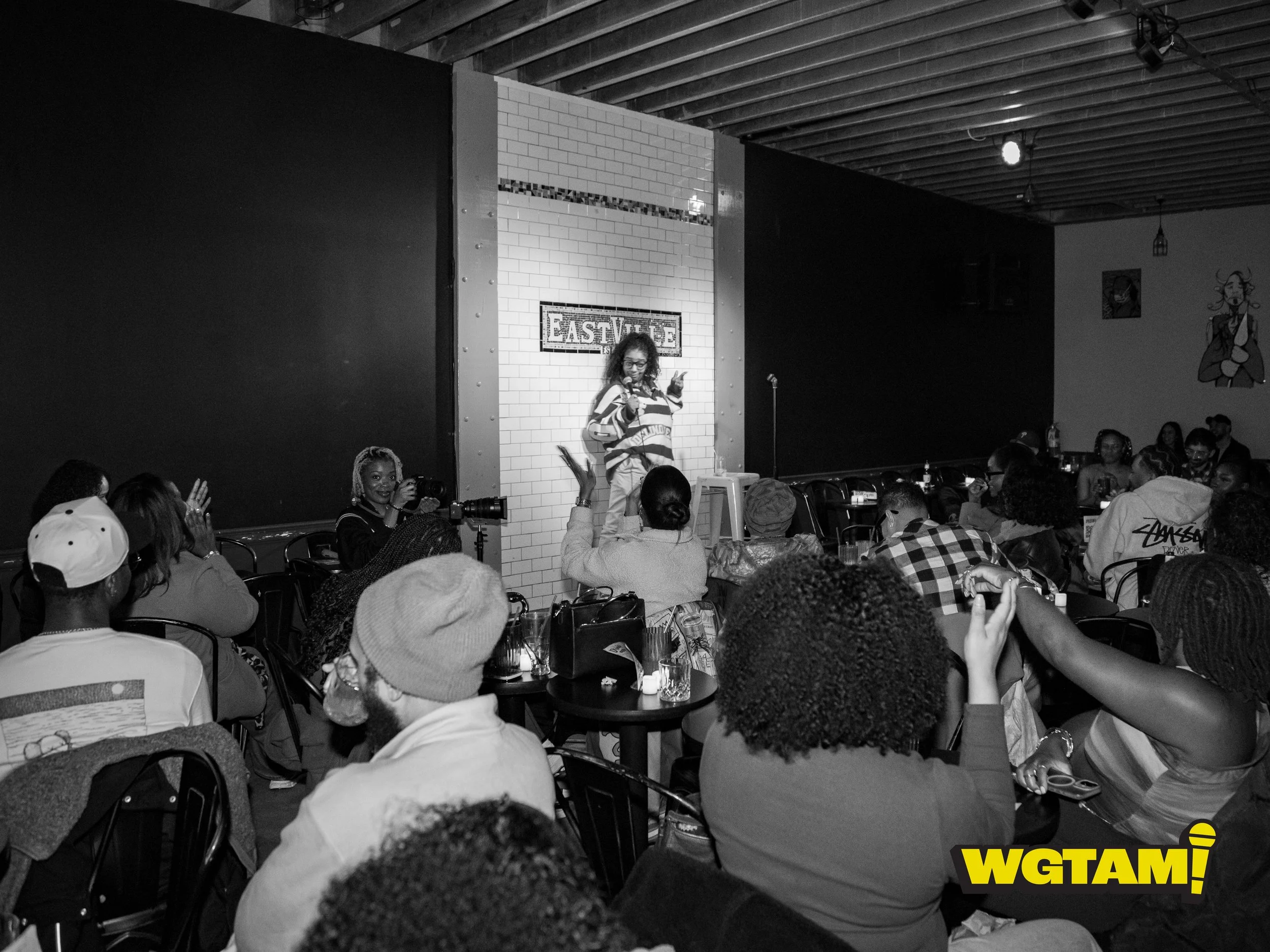 A woman performing stand-up comedy on stage at EastVille comedy club with an audience watching. The audience includes diverse individuals, some taking photos or videos, and some engaging with the performer.