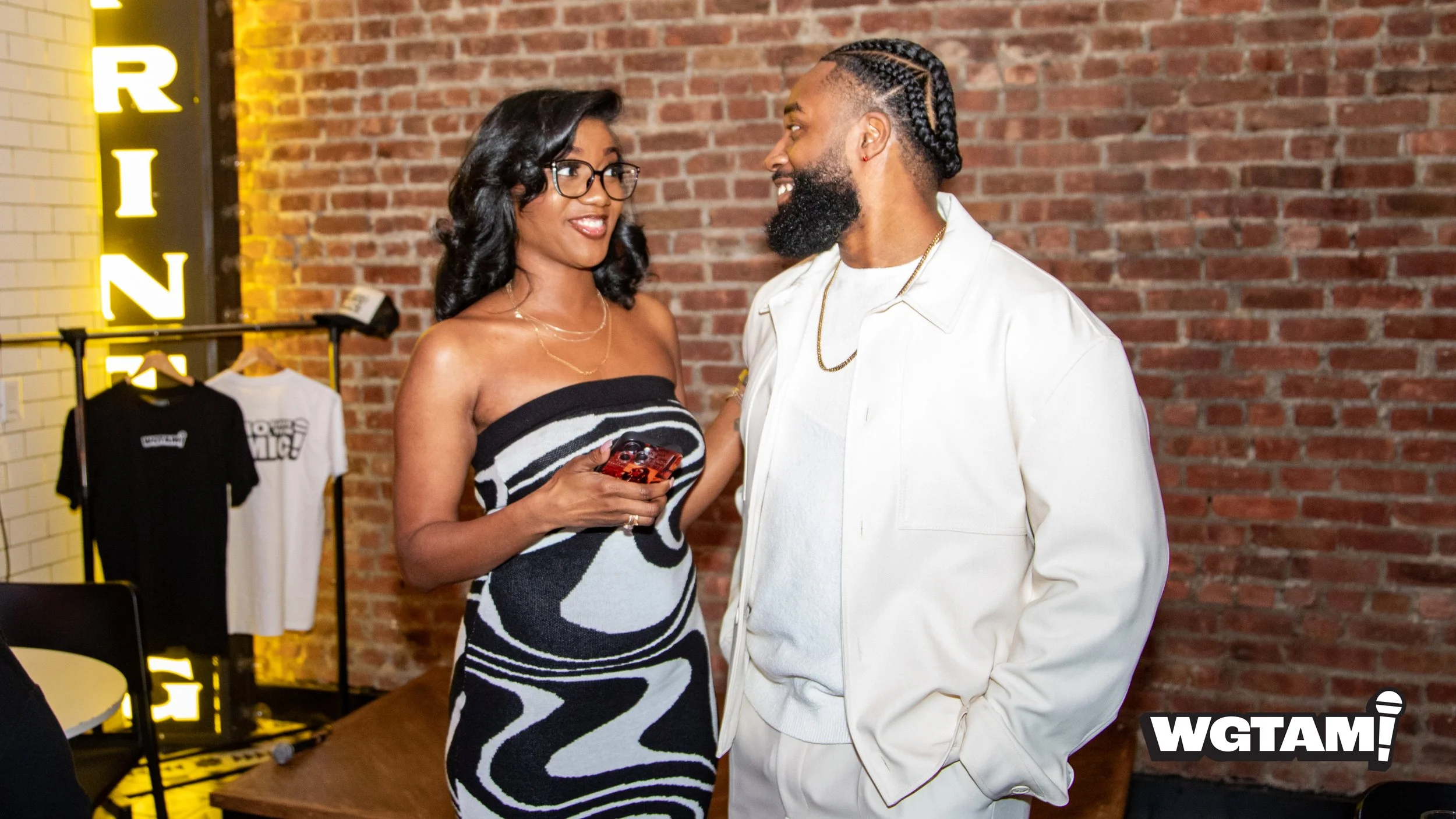 A woman and a man are standing close together, smiling and engaging in conversation indoors with a brick wall background. The woman has black hair, glasses, and is wearing a strapless black and white patterned dress, holding a phone. The man has a beard, braided hair, and is wearing a white jacket over a white shirt, with gold chains. Behind them, there is a clothing rack with shirts and a yellow illuminated sign that says 'RING.'