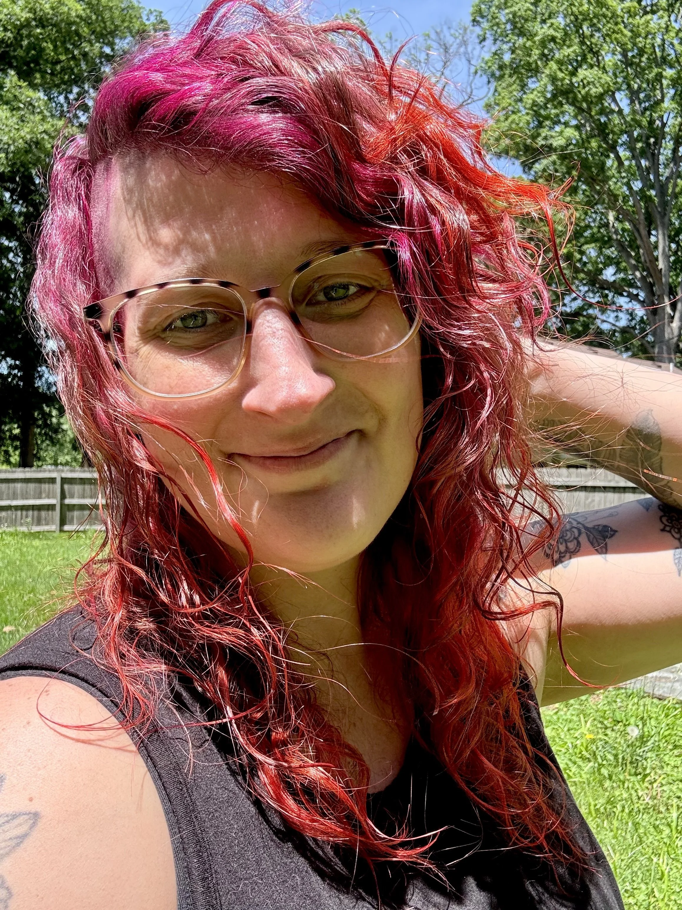 Close-up of a woman with curly red hair wearing glasses, smiling outdoors with a grassy yard and trees in the background.