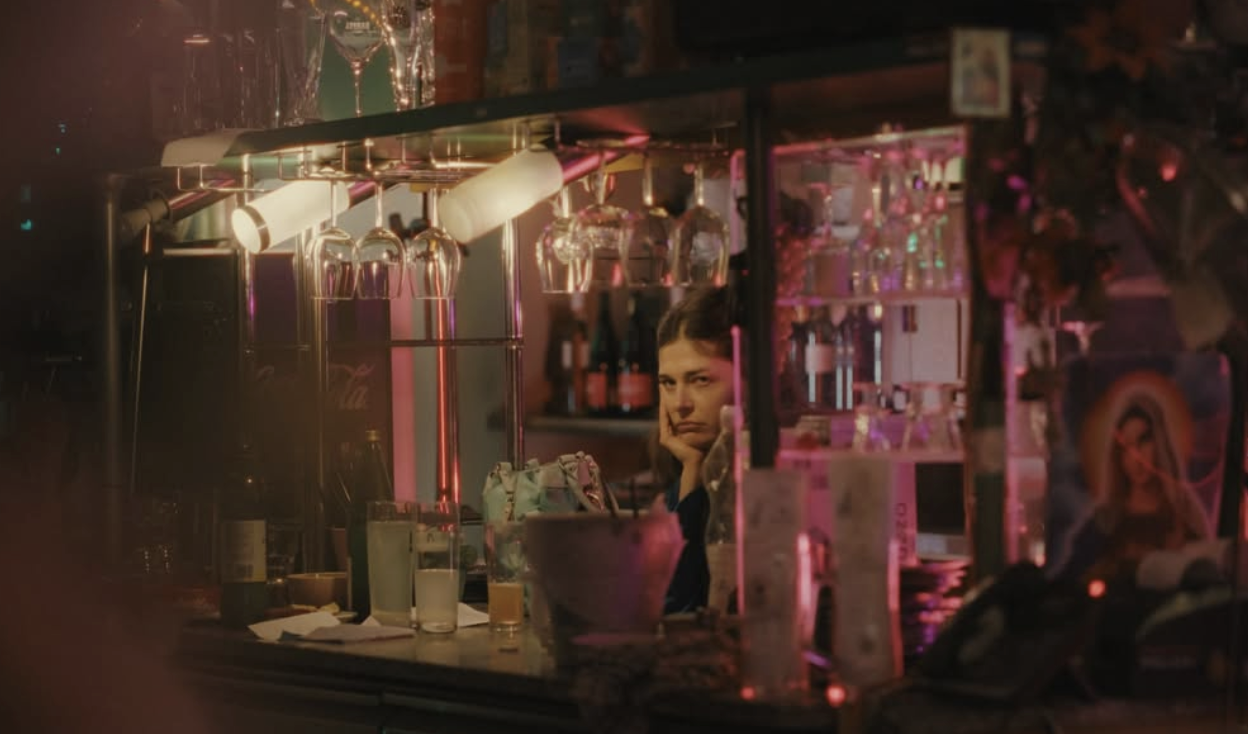 A woman sitting inside a dimly lit bar or cafe, seen through a window reflection, with various bottles and glasses on shelves behind the counter, illuminated by pink and warm lighting.
