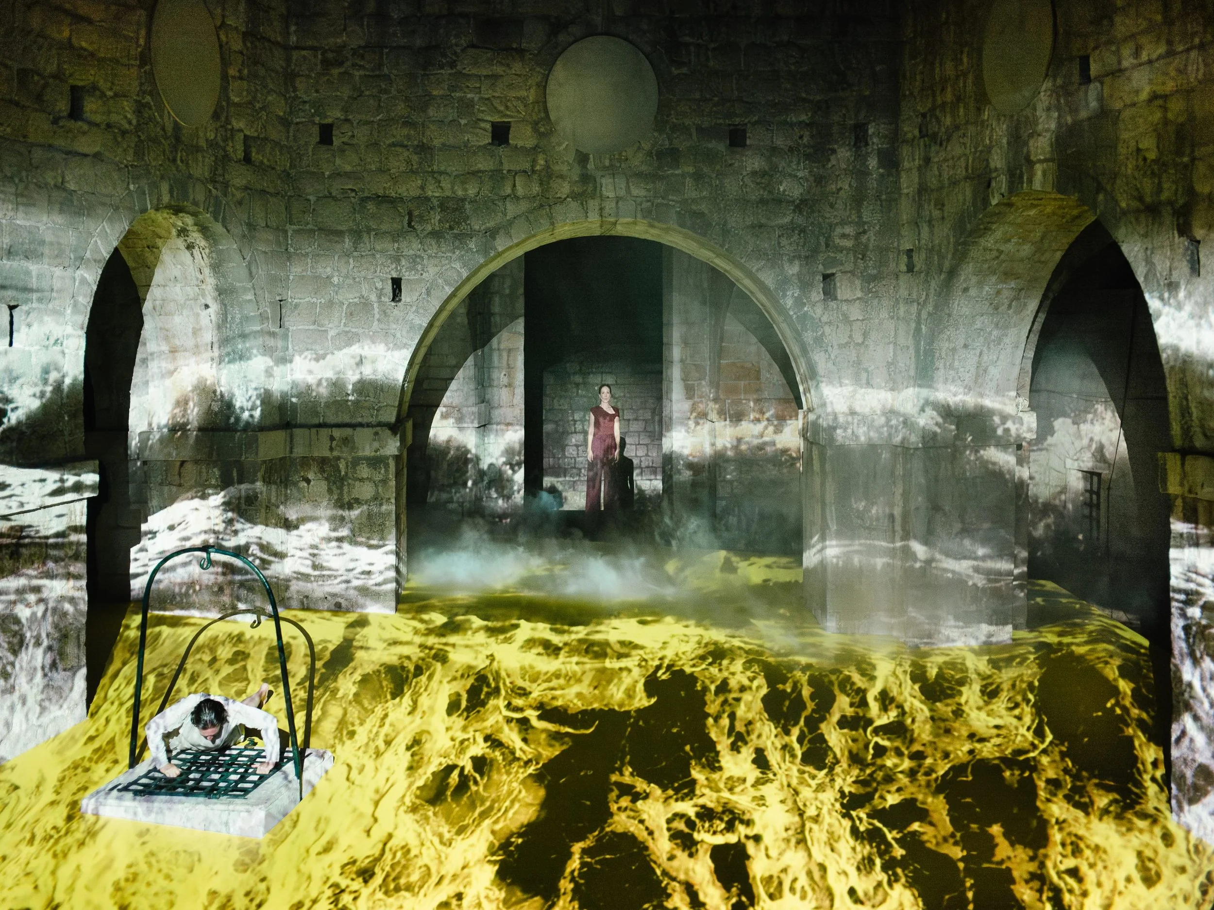An art installation featuring a person on a swing over a floor with a yellow and black marbled pattern, a brick archway, and a woman in a red dress standing in a darkened doorway, with projections of water on the walls.