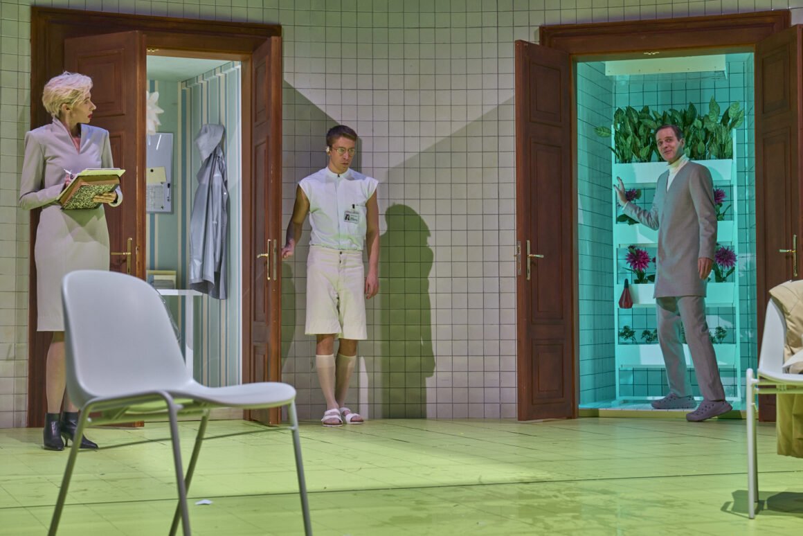 A scene from a play or movie with three characters in a room with tiled walls and open doors leading to interior spaces; one woman holding notebooks, two men in suits, one in a white vest and the other in a light gray suit, standing in separate doorways with a room of plants behind one of them.