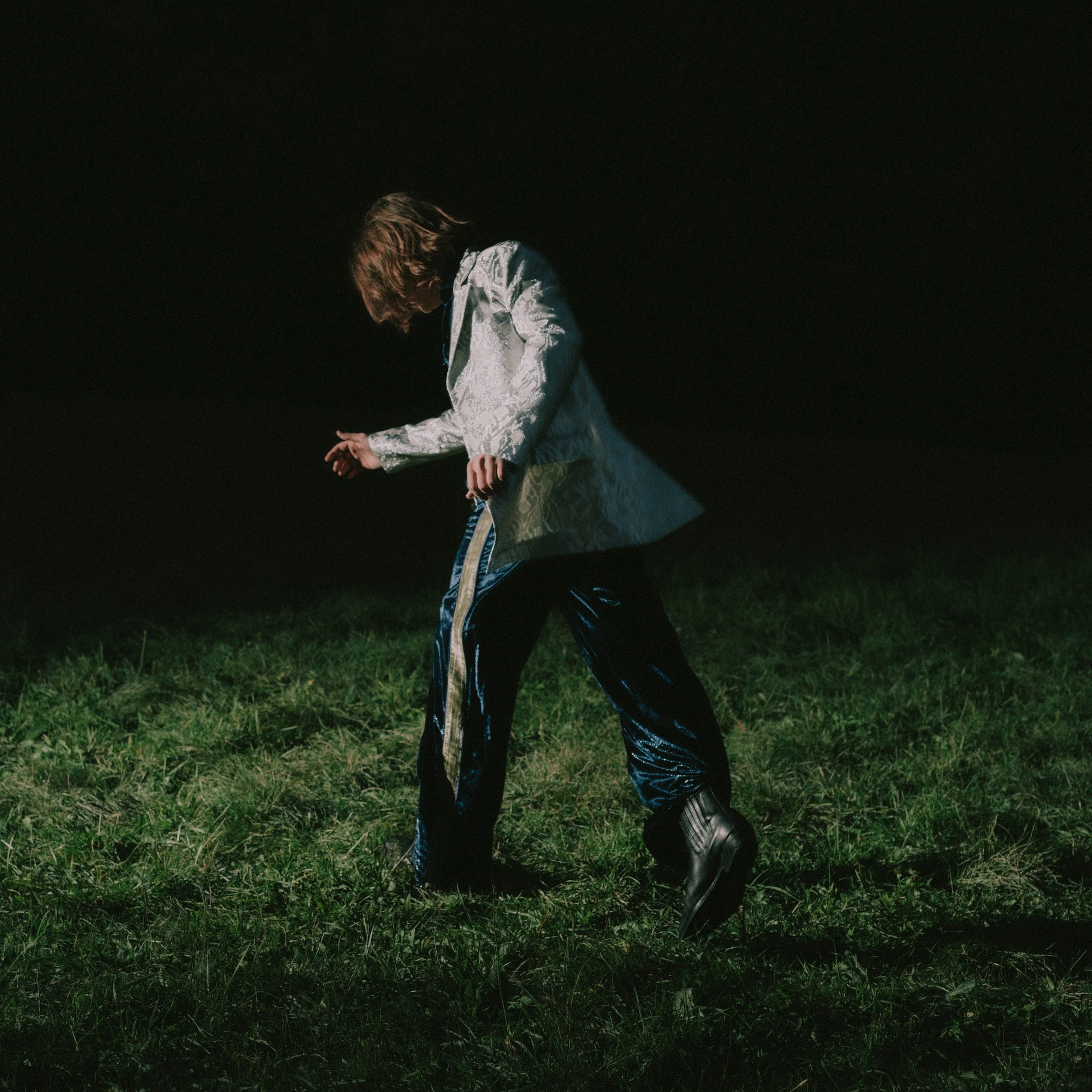 Person walking outdoors at night on grassy field, wearing a shiny silver jacket, dark blue shiny pants, and black boots.