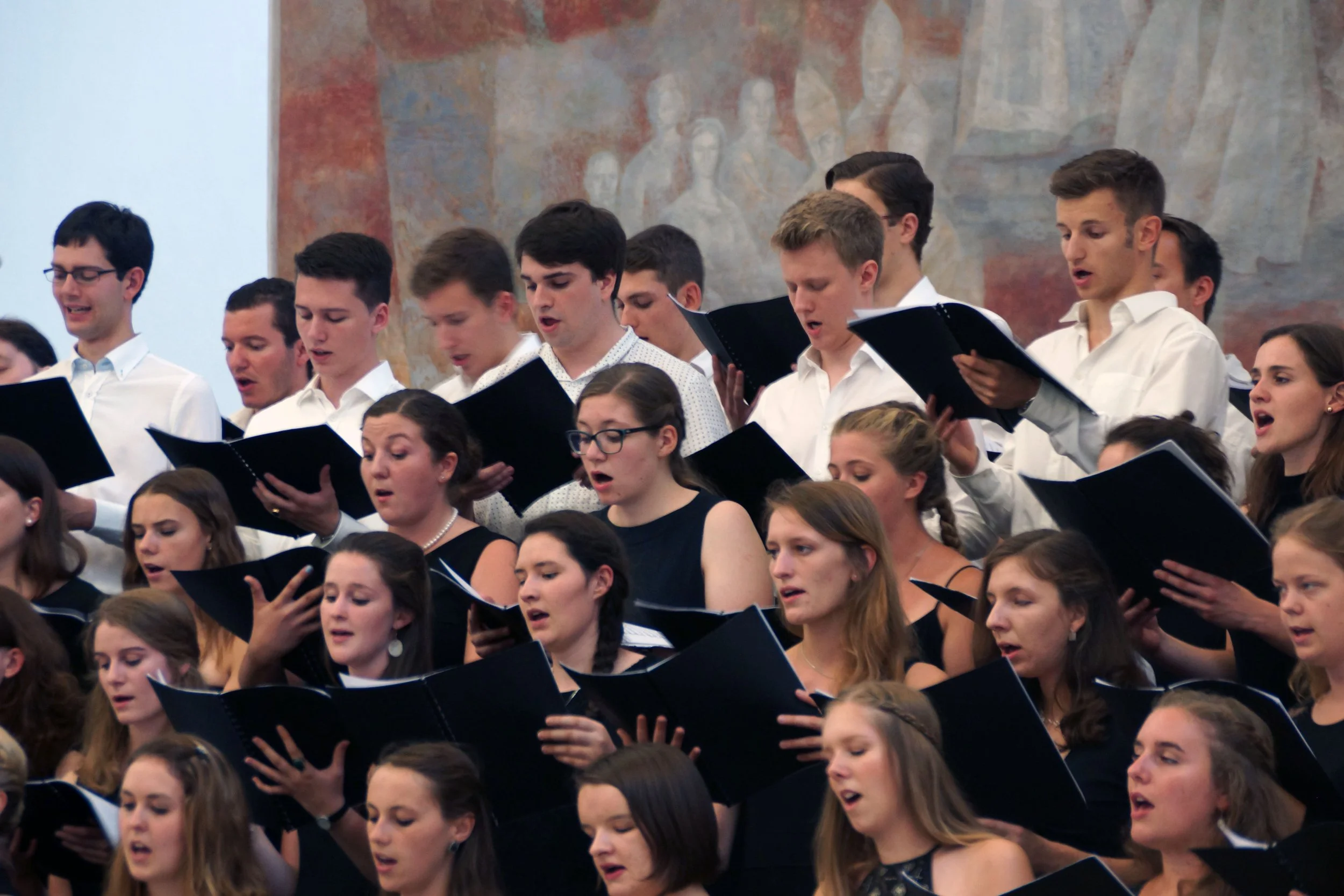 A group of young men and women singing in a choir, holding black songbooks, inside a large hall or church with a mural on the wall behind them.