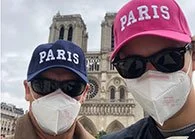 Two people wearing masks and sunglasses, each with a cap labeled 'Paris', standing in front of a historic building, likely in Paris.
