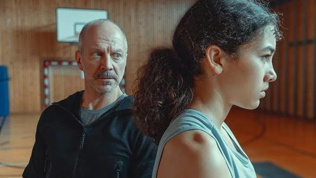 A man and a woman are sitting back-to-back in a gymnasium, with the man looking at the woman who has a serious expression.