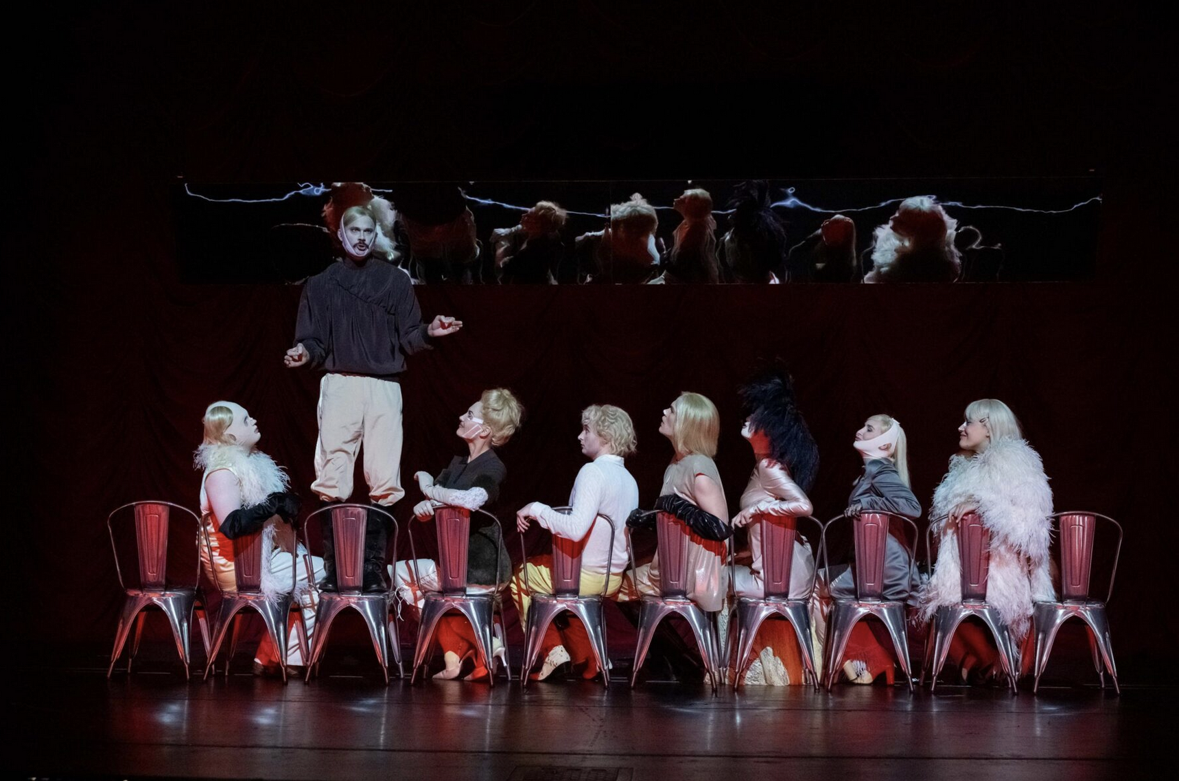 The image depicts a theatrical performance with a woman standing on a podium above a seated group of people. The seated individuals are on chairs with white and orange costumes, and some wear feathered accessories and gloves. The stage has a dark background with a mirror or reflective surface above, reflecting the scene below. The lighting highlights the performers, and the scene suggests a dramatic or artistic presentation.