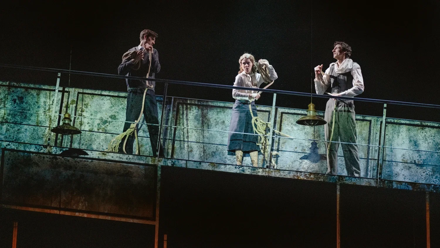Three actors on a stage dressed in period costumes, engaged in a theatrical scene with a weathered backdrop.