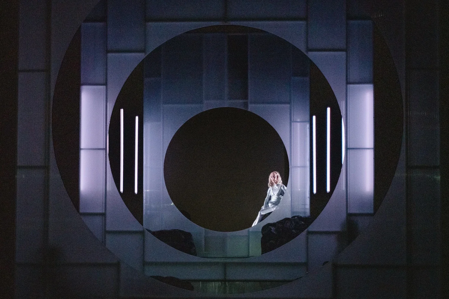 A woman sitting on a ledge within a futuristic, circular metallic structure with glowing vertical lights.