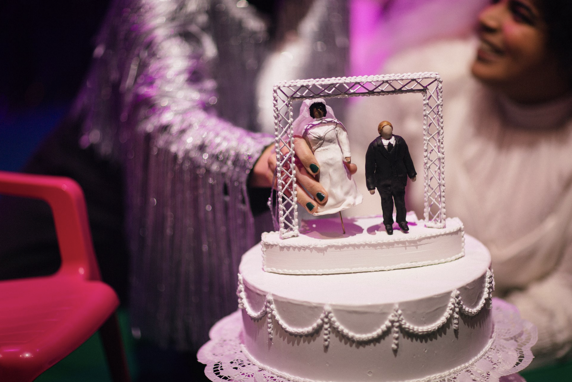 A wedding cake with a topper featuring miniature bride and groom figurines, one dressed as a bride in a white dress and the other as a groom in a black tuxedo, decorated with white icing and intricate piping, with a person in a shiny, textured silver jacket in the background.