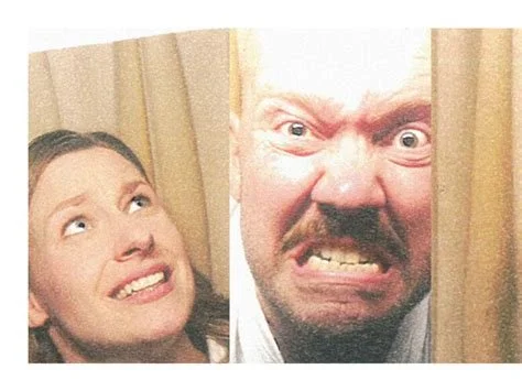 A woman and a man press their faces against a curtain, making exaggerated facial expressions of amusement and anger.