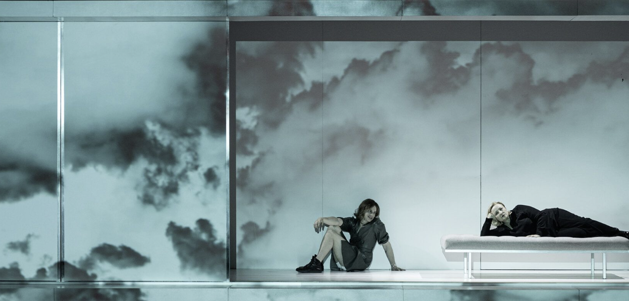 Two women in a modern room with cloud-patterned wallpaper and glass walls, one sitting on the floor and the other lying on a bed, both looking at each other.