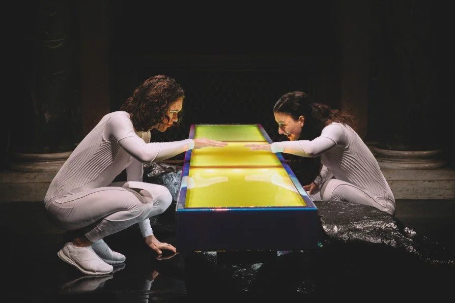 Two women in white outfits and glasses are sitting cross-legged on the floor, leaning over a brightly lit, colorful table with their hands touching its surface, creating a mirror-like reflection of each other.