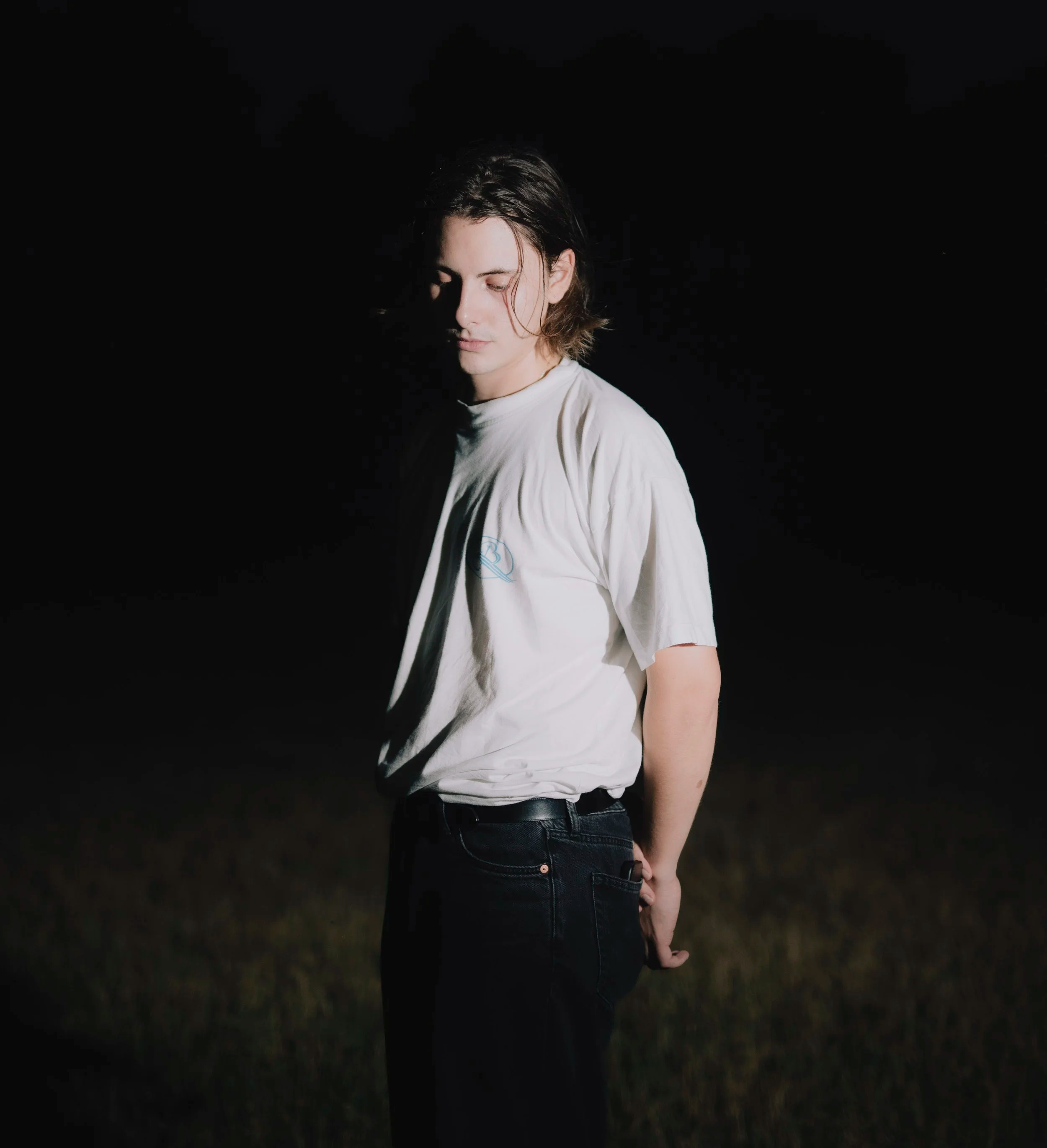 Aki Traar, Producer, Music, Composer. A young person with medium-length brown hair stands with closed eyes in a dark outdoor setting, wearing a white t-shirt and black jeans.