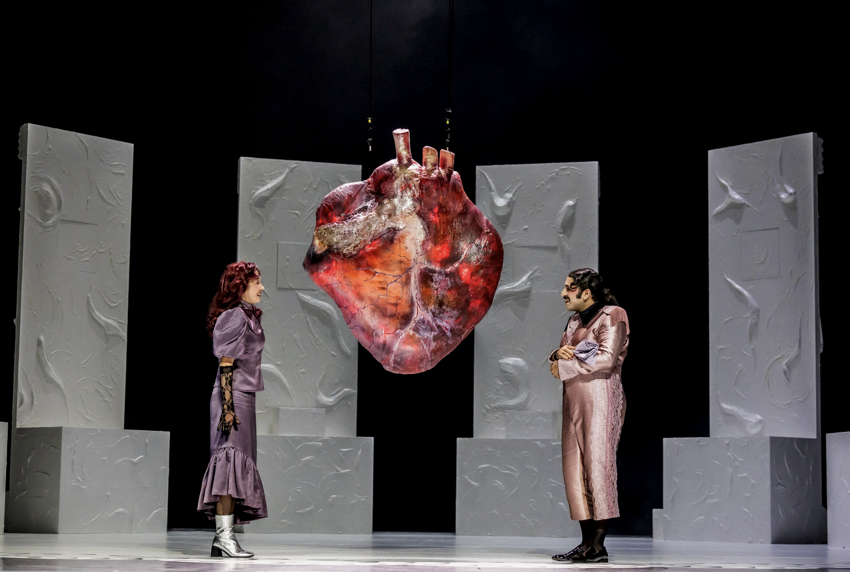 Two performers, one woman with wavy red hair in a purple dress and another with dark hair in a pink outfit, stand on stage facing each other. They are surrounded by white textured panels and a hanging prop shaped like a large human heart.
