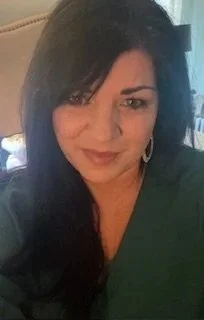 A woman with dark hair in a side part, wearing hoop earrings and a green top, smiling slightly in an indoor setting.