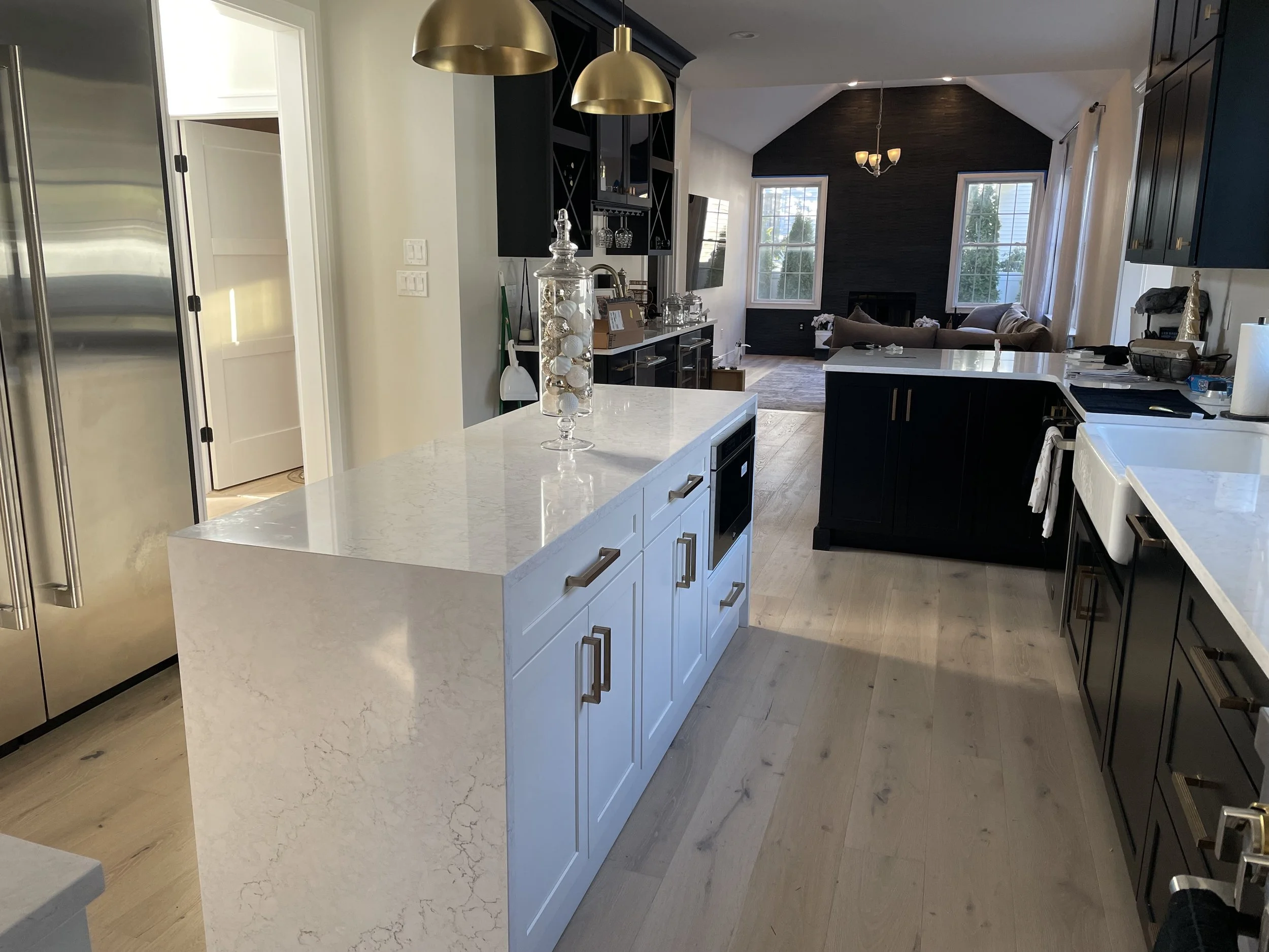 Modern kitchen with white island and black cabinets, stainless steel refrigerator, and light wood flooring, with a living room visible in the background.