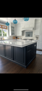 Modern kitchen with white cabinets, a large white island with a dark blue base, and blue pendant lights hanging above. Contains a window, some small appliances, and a decorative backsplash.