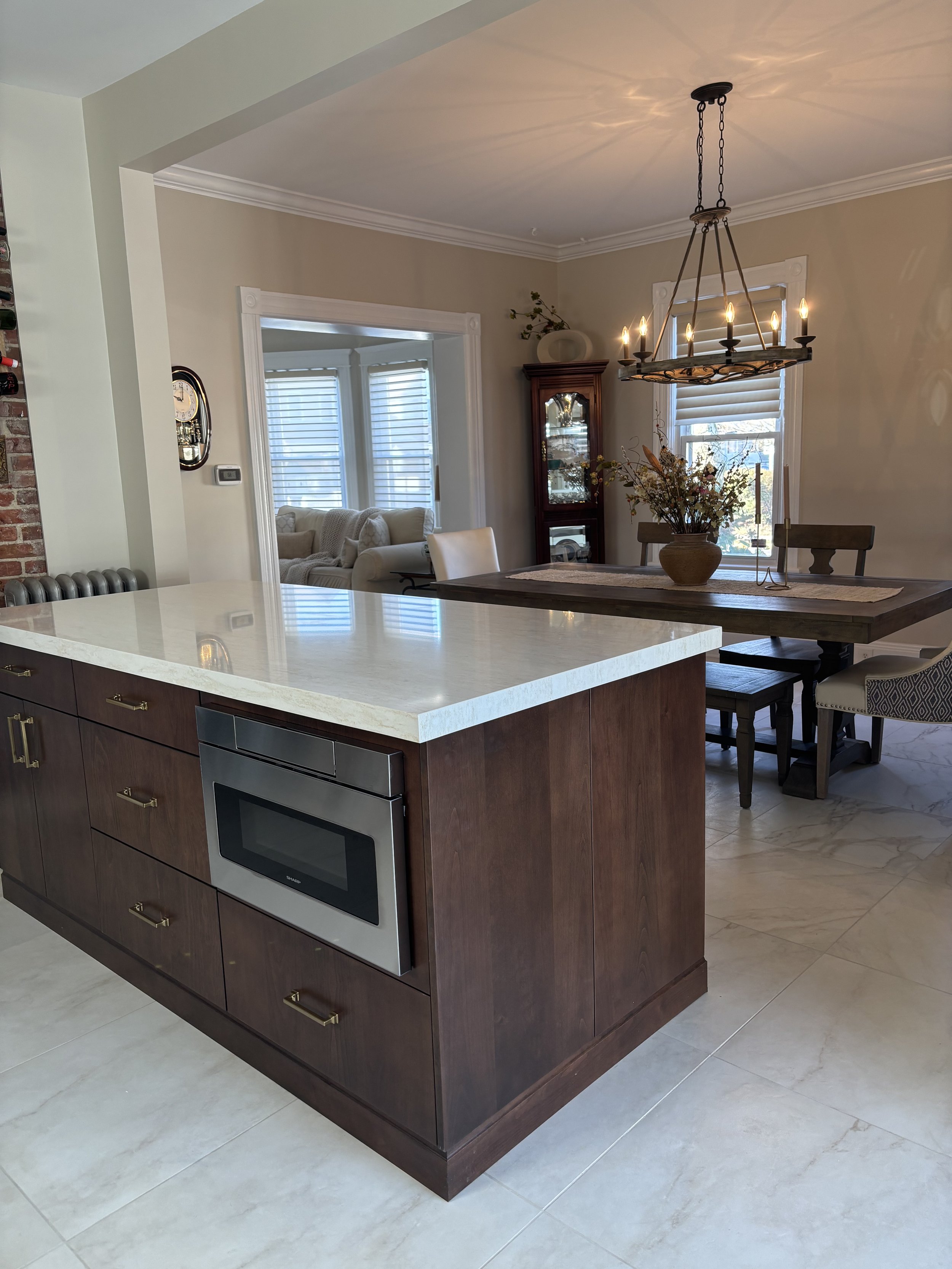 Kitchen island with a built-in microwave, dark wooden cabinetry, white marble countertop, and a dining area with a chandelier, wooden table, chairs, and a vase with flowers.