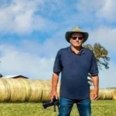 Glenn Johnson, photographer, wearing a hat and sunglasses, standing in a field with large hay bales and a tree in the background.