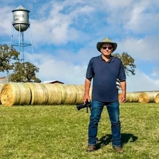 Man standing on grass field in front of hay bales, holding camera, wearing hat and sunglasses, blue shirt, with water tower in background under a partly cloudy sky.