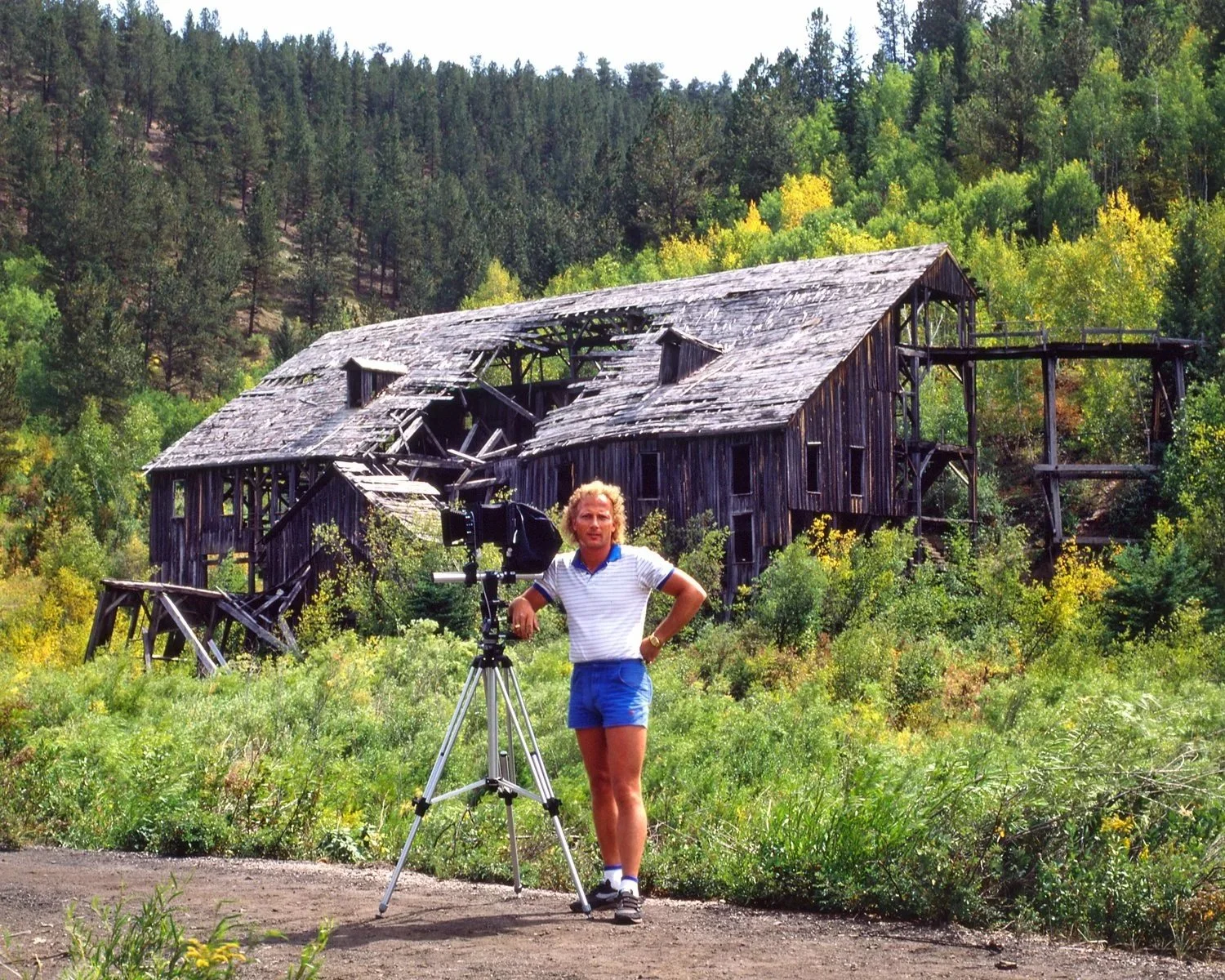 Glenn Johnson, back in the day, early in his career as a professional photographer, curly blonde hair, striped shirt, and shorts stands next to a vintage camera on a tripod in front of a dilapidated wooden building in a lush forested area.