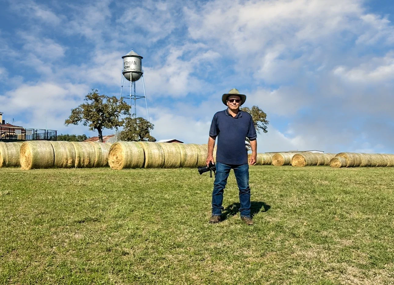 Glenn Johnson, photographer, standing on grass holding a camera, wearing a hat and sunglasses, with hay bales in the background and a water tower labeled 'NATHAN' under a partly cloudy sky.