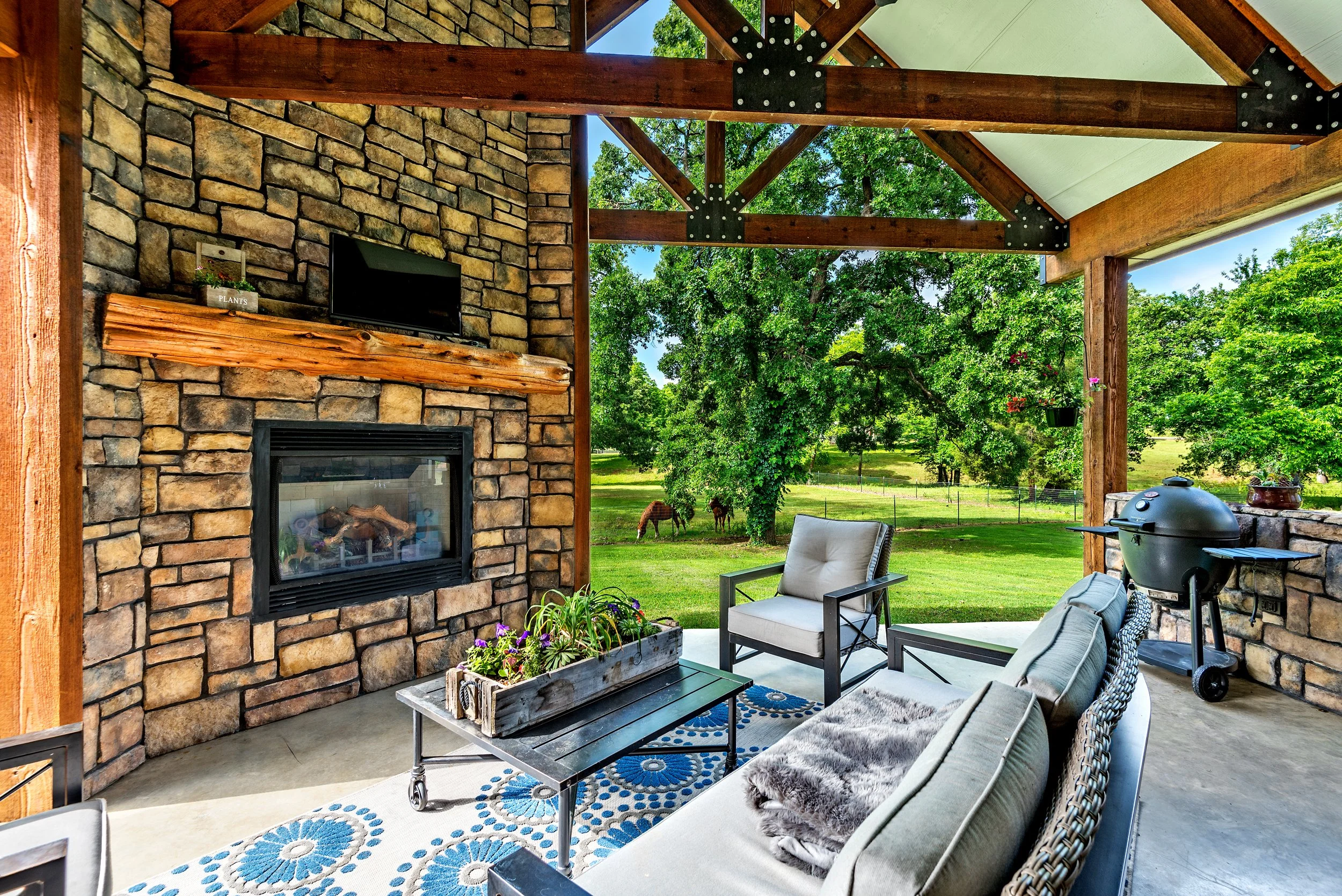 Covered outdoor seating area with stone fireplace and a mounted TV, overlooking a grassy yard with trees and grazing horses.