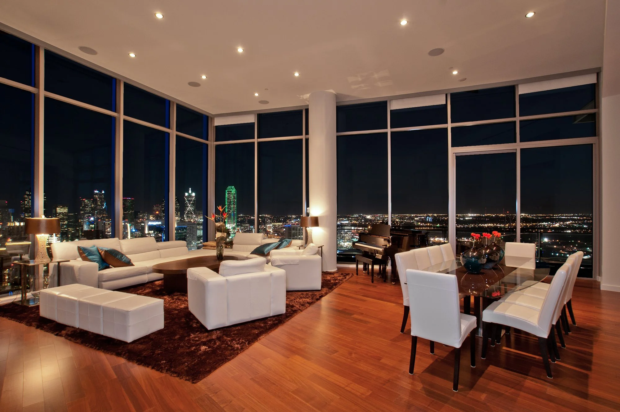 Modern high-rise living room with large floor-to-ceiling windows showing a city skyline at night, white furniture including sofas and chairs, a round wooden coffee table, and a dining table with white chairs, illuminated by warm ambient lighting.
