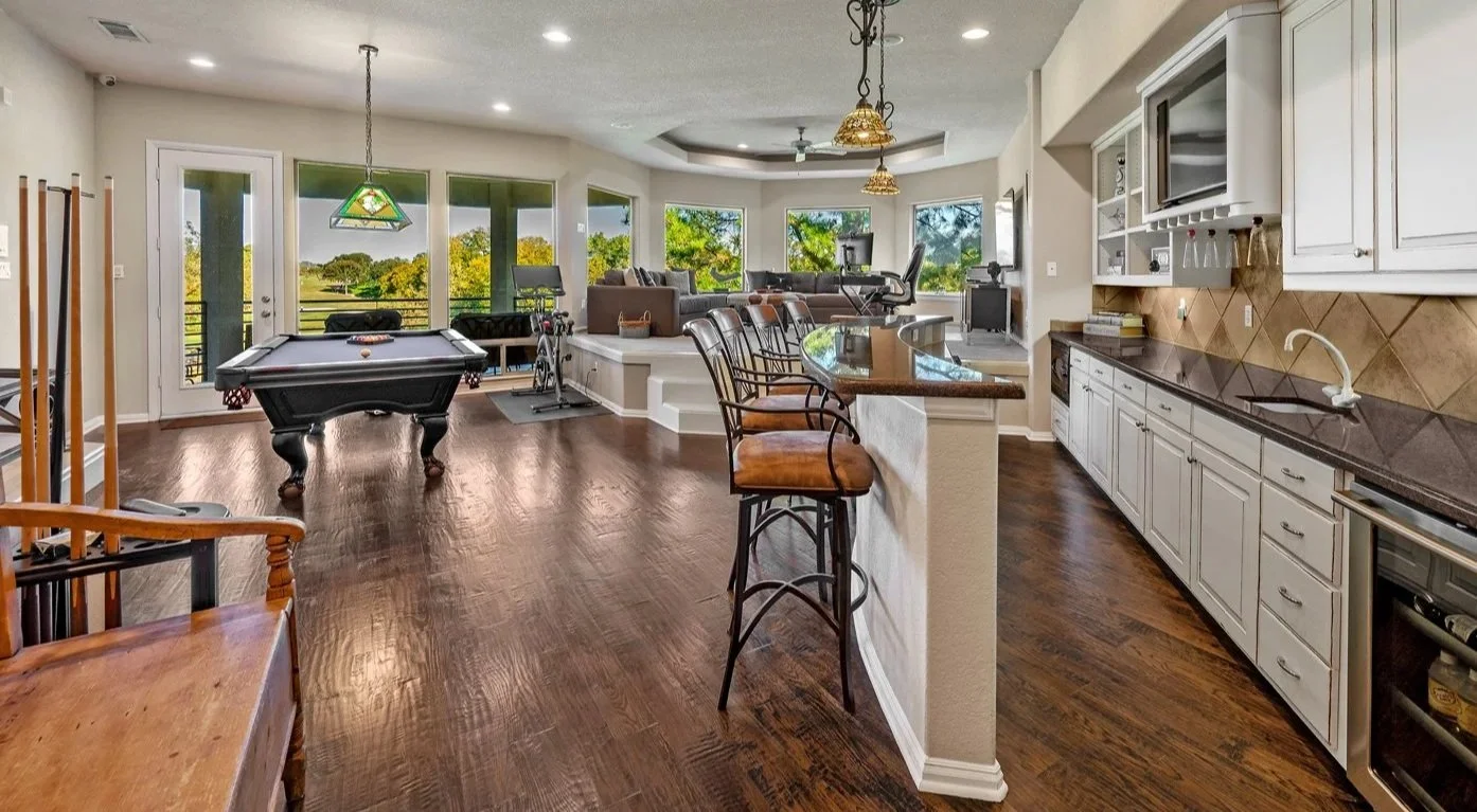 Spacious open-concept living area with kitchen, bar counter, recreational space, pool table, and multiple seating areas, overlooking a scenic outdoor landscape through large windows.