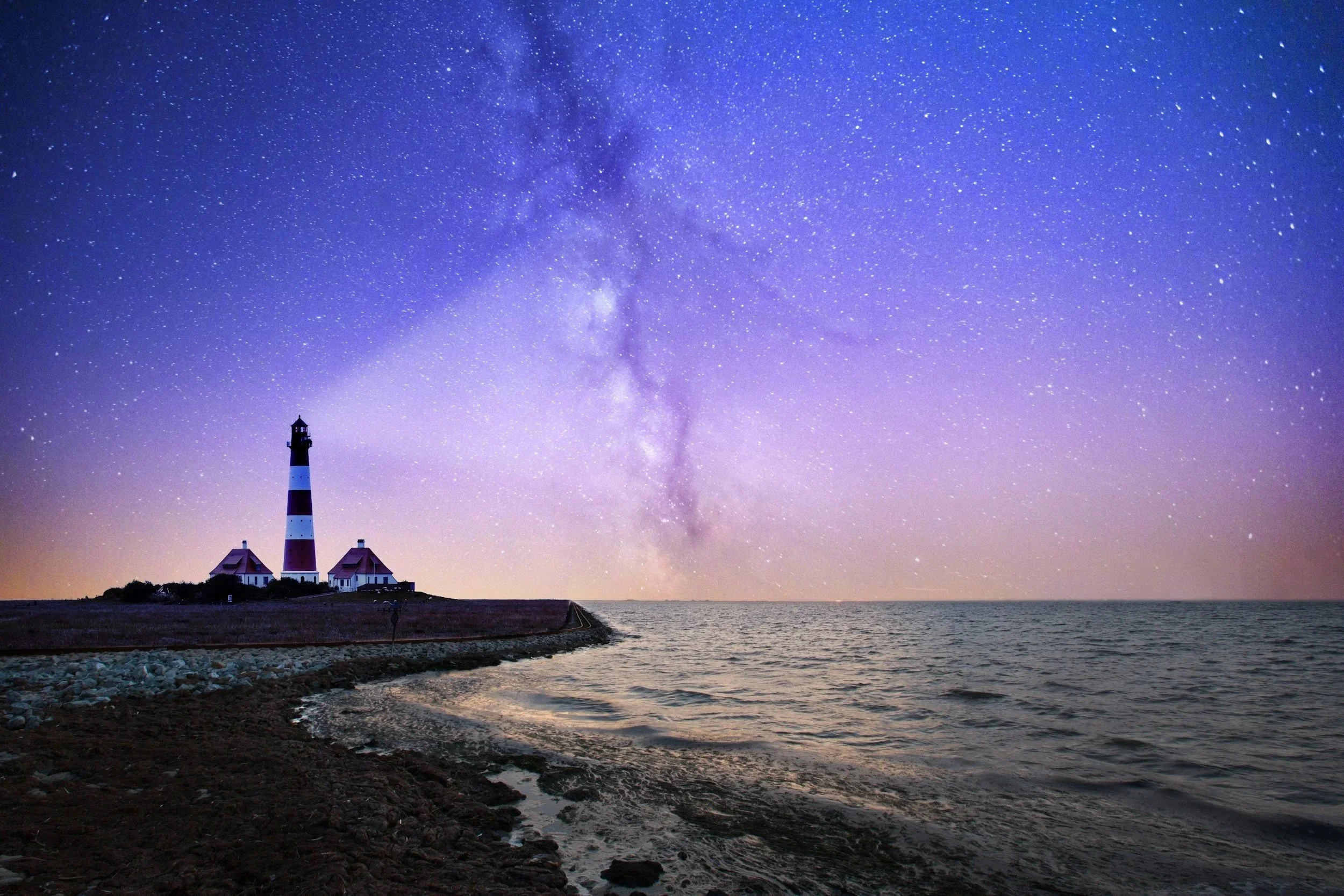 A lighthouse under a starry night sky with the Milky Way visible across the sea.