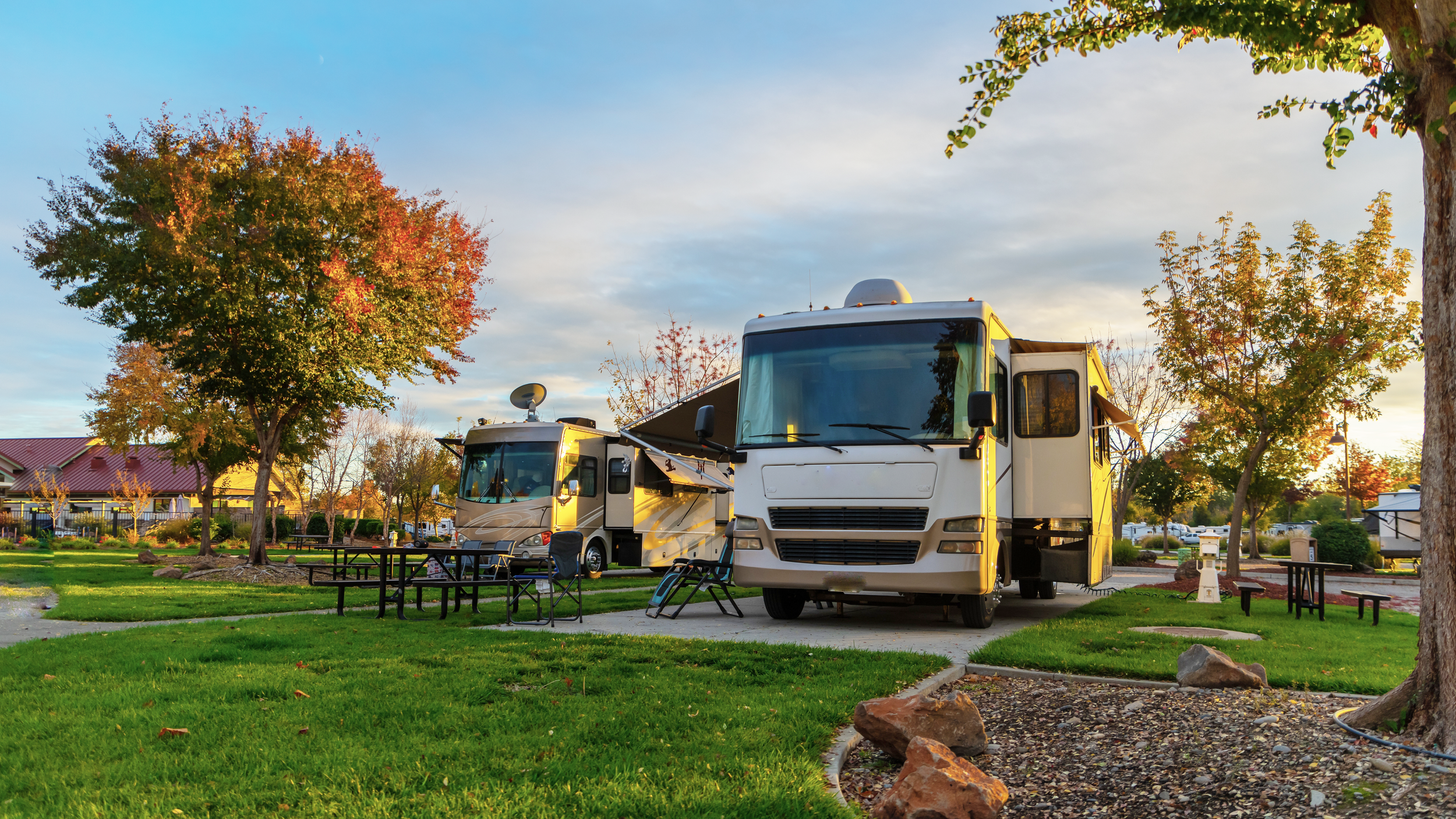 Two motorhomes parked at a campsite with trees showcasing autumn colors, outdoor chairs, and tables in a well-maintained grassy area.